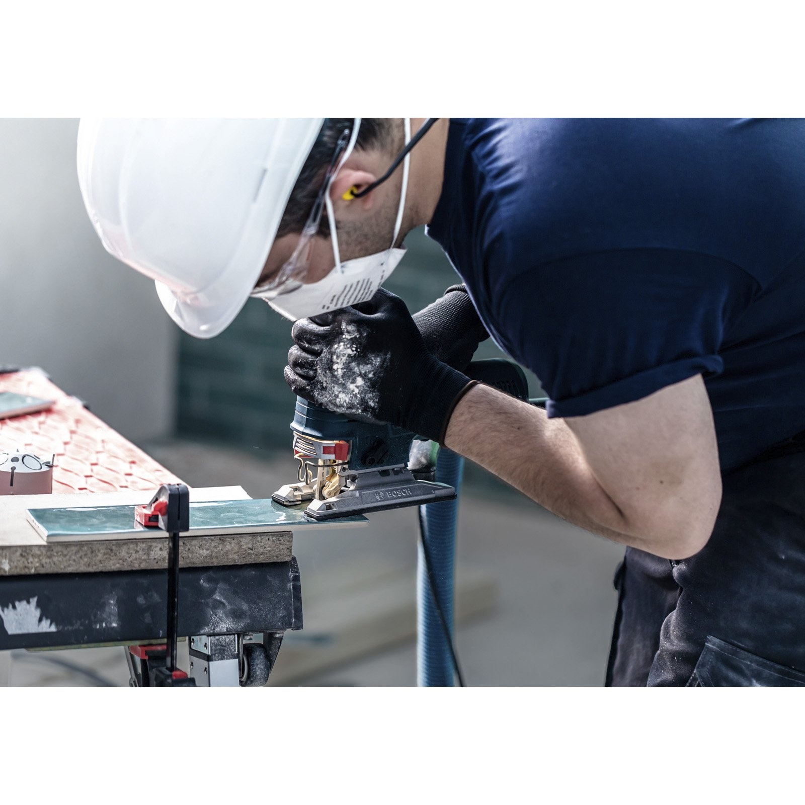 Pracownik w sprzęcie ochronnym przecina płytki za pomocą wyrzynarki Bosch na stole warsztatowym.