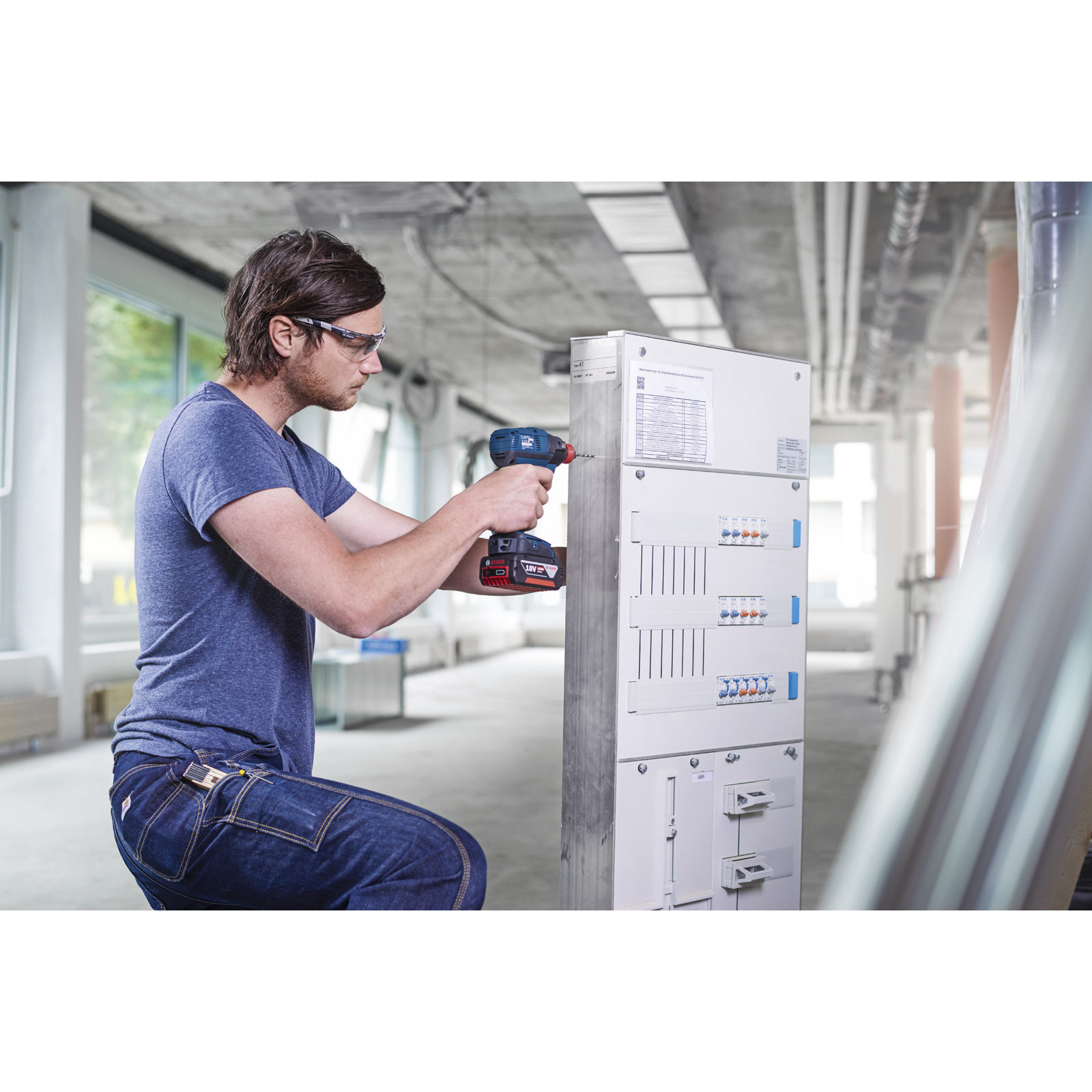 Mann in blauem Hemd und Jeans, mit Schutzbrille, bohrt in einem industriellen Umfeld in eine Schalttafel.