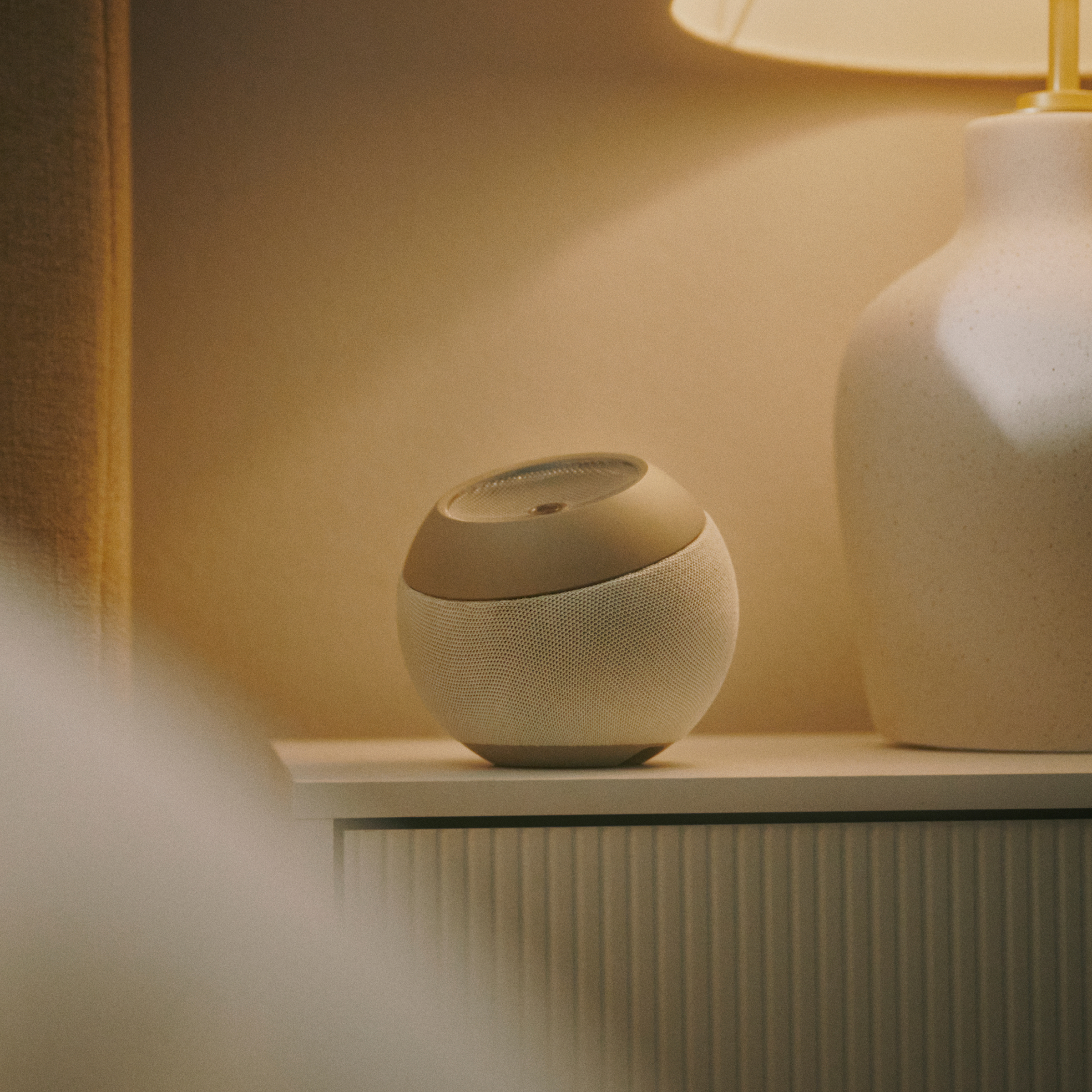 Runder Lautsprecher auf einem Nachttisch neben einer Lampe. Der Lautsprecher hat eine Stoffstruktur.