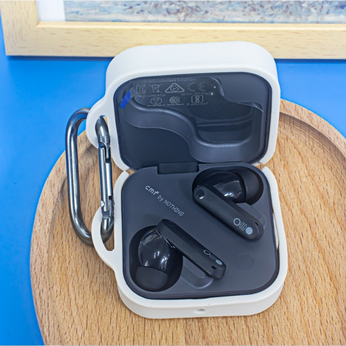 Auricolari in una custodia bianca quadrata con clip in metallo, su una superficie di legno su sfondo blu.