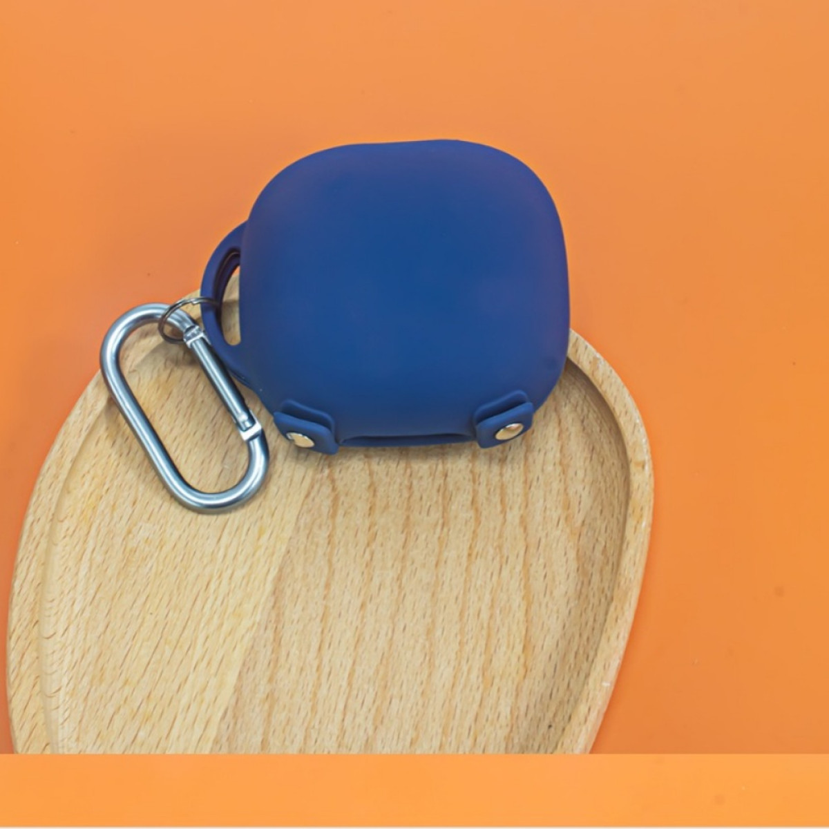 Blaues Ohrhörer-Etui mit silbernem Karabinerhaken auf einem Holztablett, vor orangefarbenem Hintergrund.