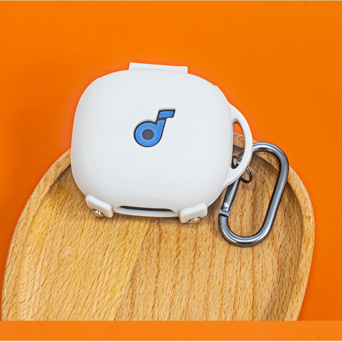 Weißes Ohrhörer-Etui mit blauem Logo, silbernem Karabiner, auf Holztablett vor orangefarbenem Hintergrund.