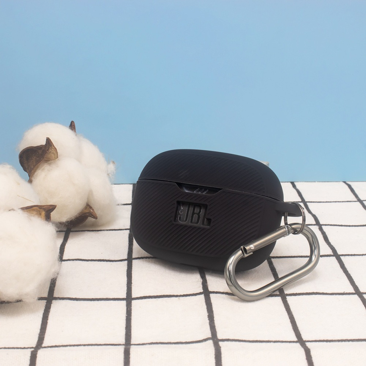 Schwarzes Ohrhörer-Etui mit JBL-Logo, silbernem Karabiner, auf gemusterter Oberfläche mit Baumwollpflanzen.
