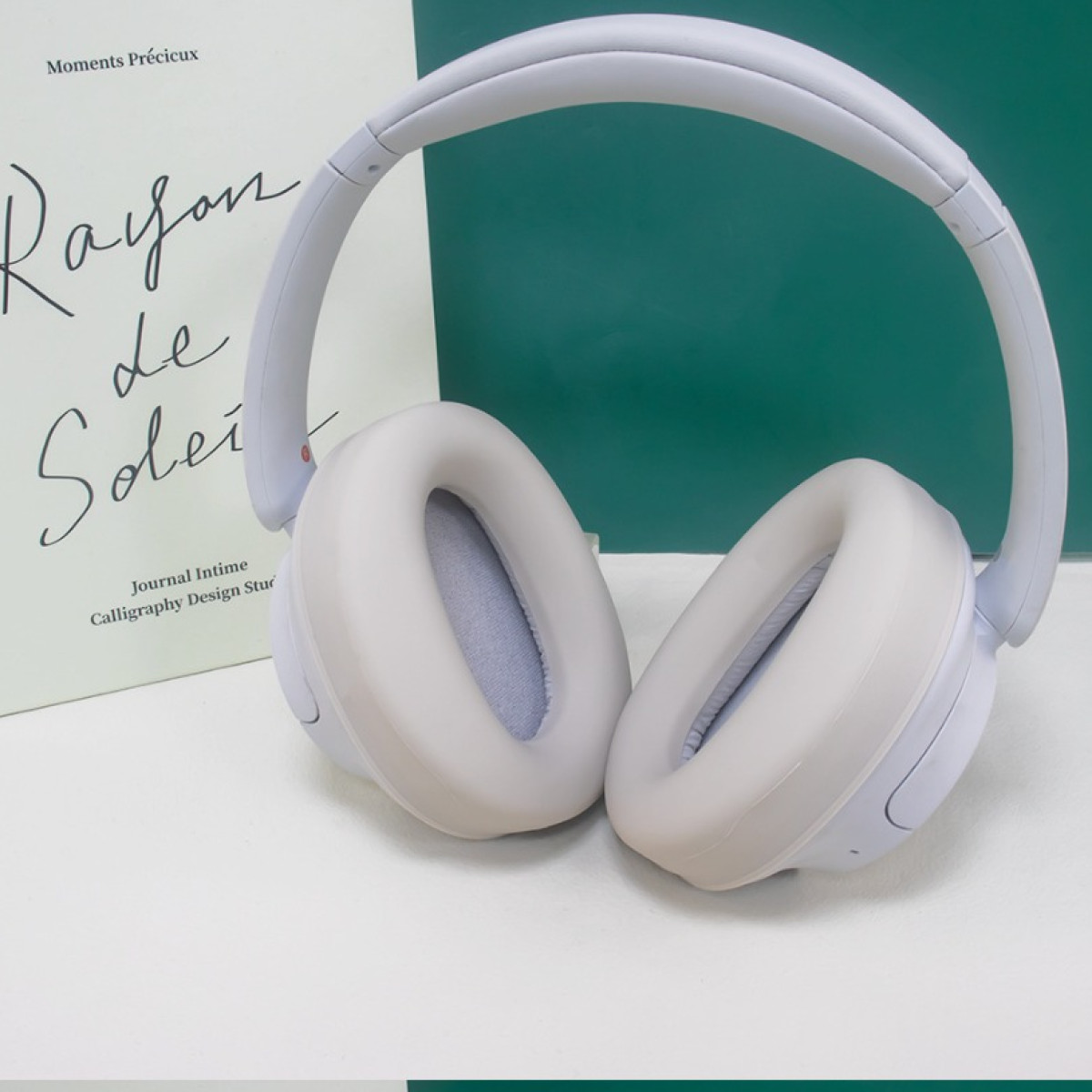 Weiße Over-Ear-Kopfhörer mit weichen Ohrmuscheln, in der Nähe eines Buches auf einer weißen Oberfläche mit einem dunkelgrünen Akzent.