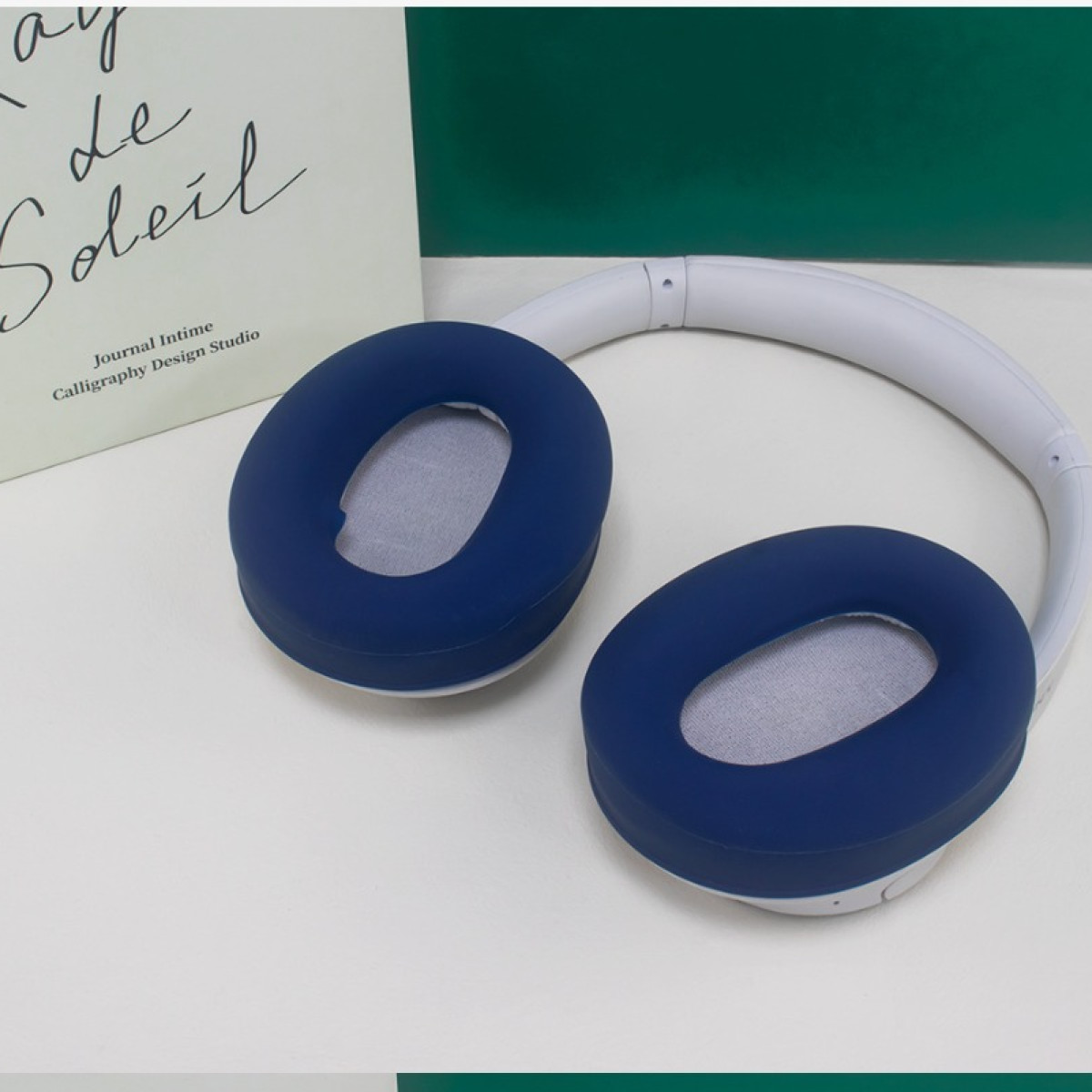 Cuffie bianche con padiglioni auricolari blu scuro vicino a un libro e un pannello verde su una superficie bianca.