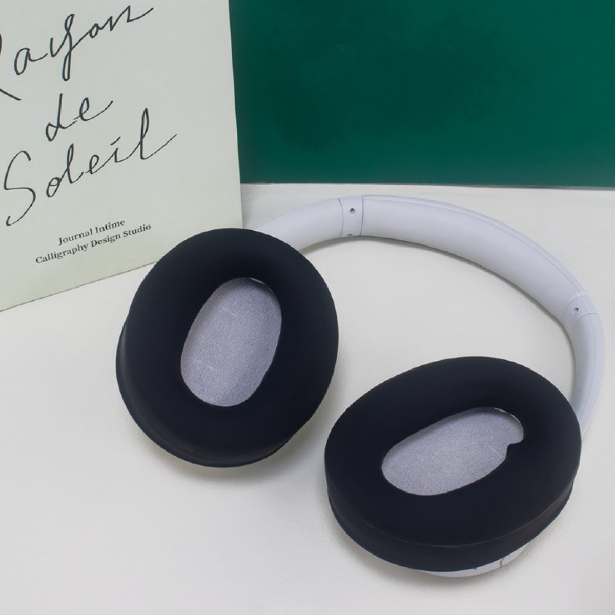 Cuscinetti auricolari neri su cuffie bianche sono posizionati accanto a un libro su una superficie bianca, contro un pannello verde.