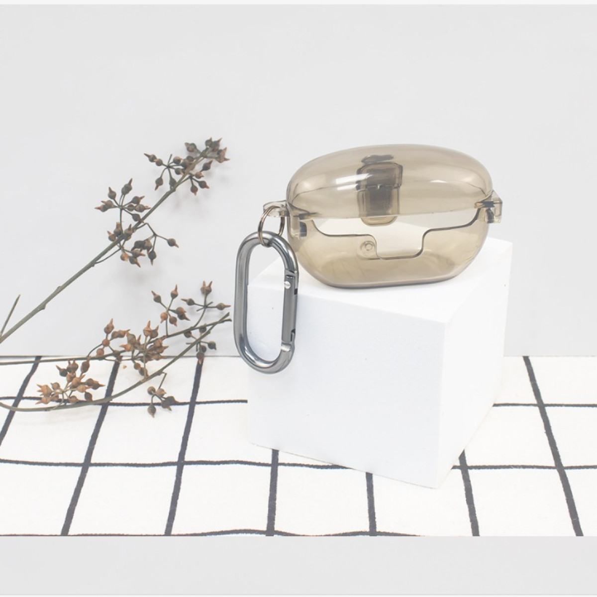 Durchsichtiges, graues Ohrhörer-Etui mit silbernem Karabiner, ruhend auf einem weißen Würfel, Zweig- und Gitterhintergrund.