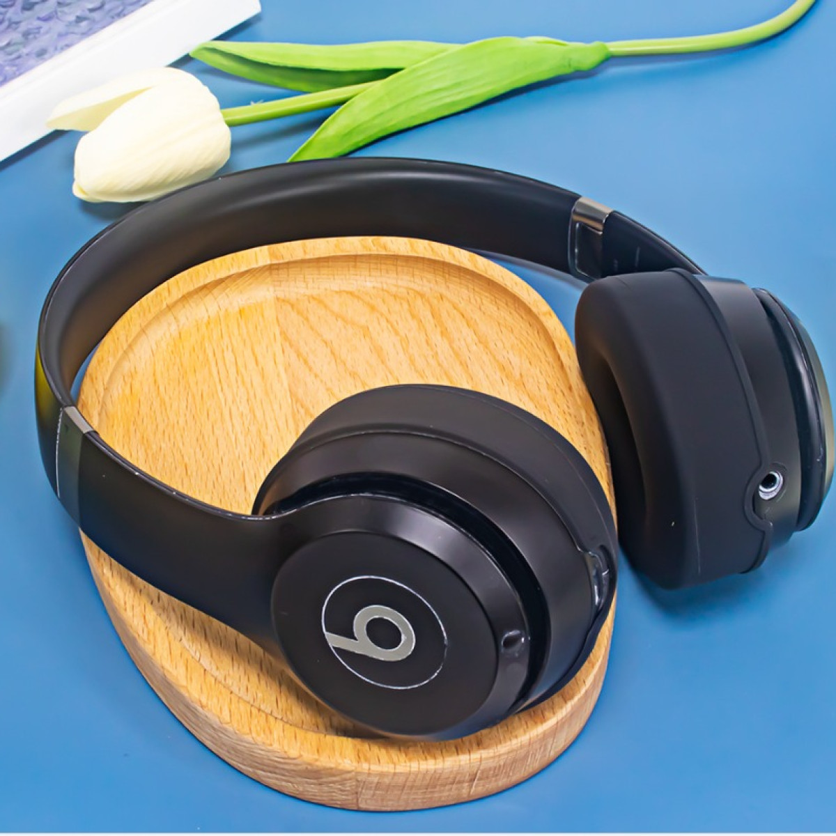 Cuffie Beats nere appoggiate su un supporto di legno con un tulipano bianco e uno sfondo blu, che mostrano accessori audio.
