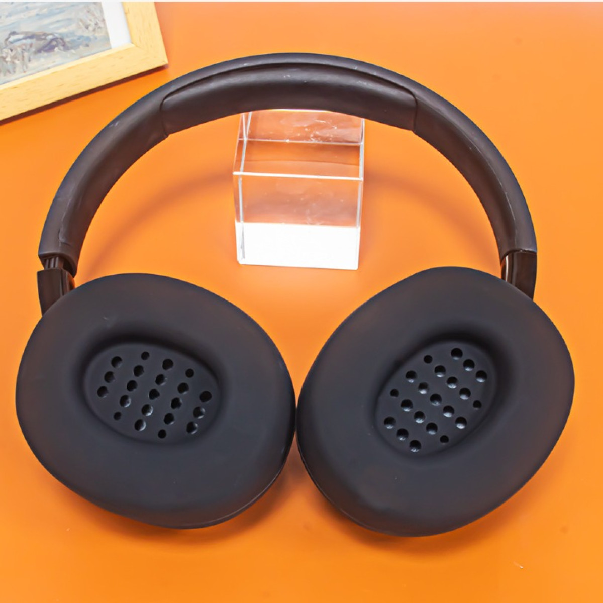 Cuffie nere con padiglioni forati sono su una superficie arancione, con un'immagine incorniciata e un cubo trasparente sullo sfondo.