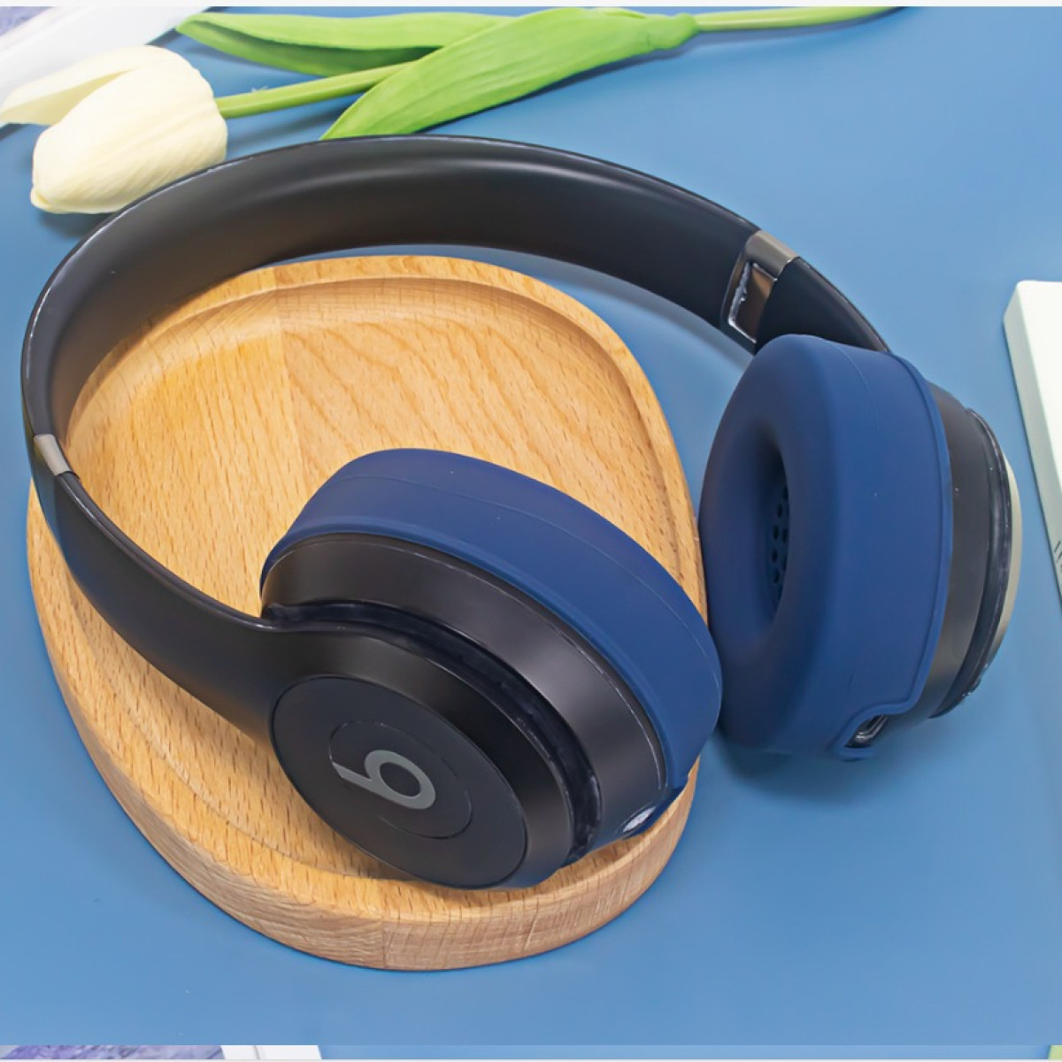 Le cuffie Beats blu e nere appoggiano su un supporto di legno, con un tulipano bianco e un libro sullo sfondo.