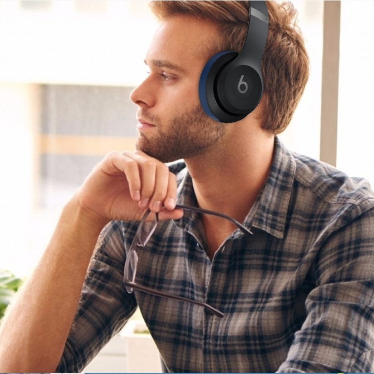Un uomo che indossa cuffie Beats nere e blu tiene in mano degli occhiali, con una camicia a quadri e uno sfondo sfocato.