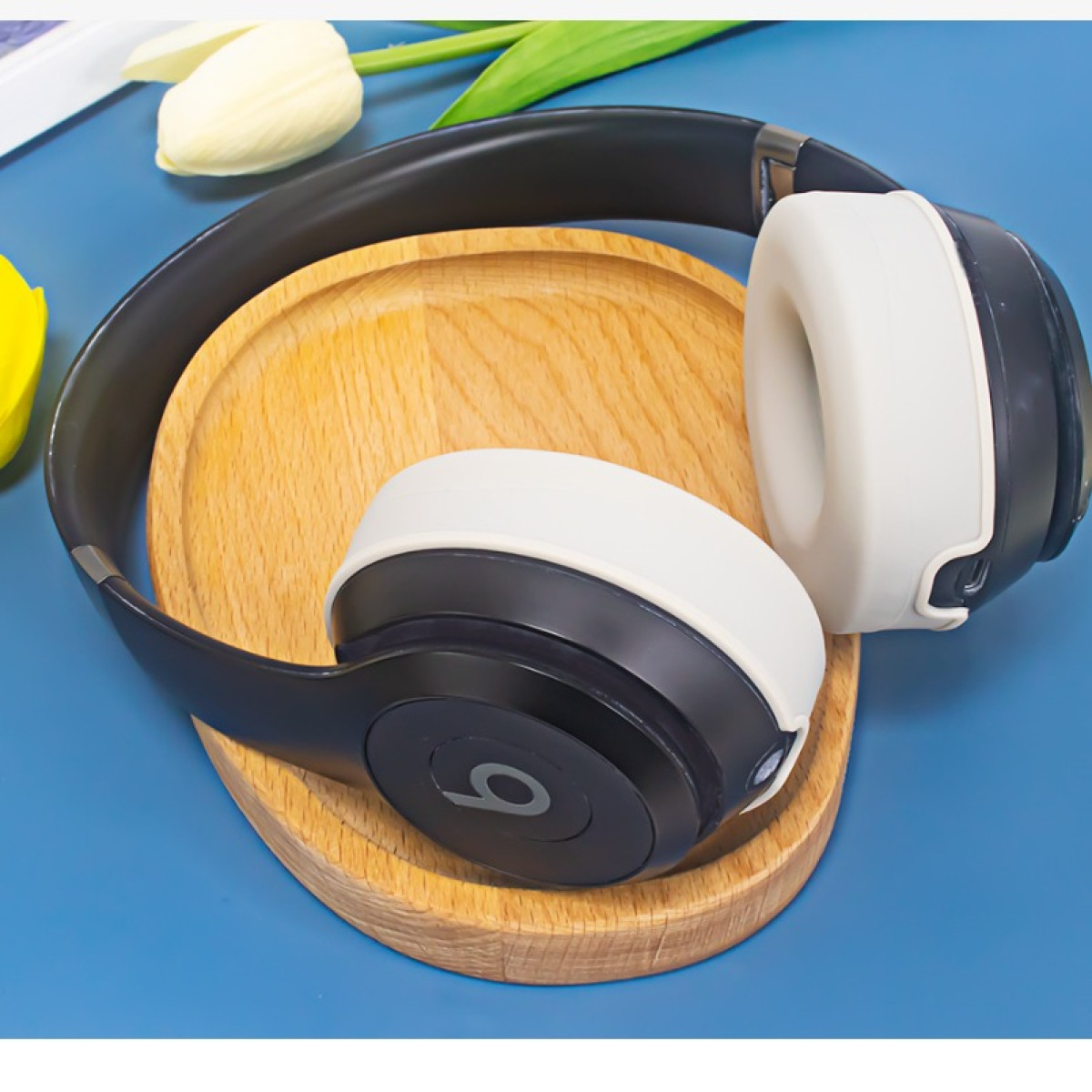 Cuffie bianche e nere su un sottobicchiere di legno, con un tulipano bianco sullo sfondo, il tutto su una superficie blu.