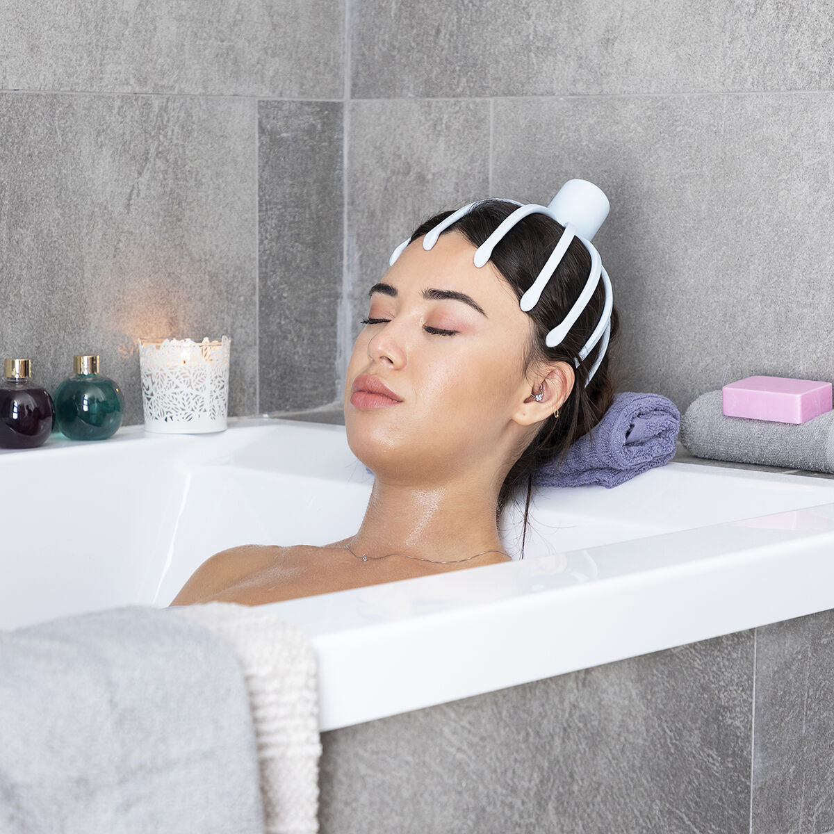 Mujer relajada en una bañera con masajeador de cabeza. Vela y productos de baño en el borde.