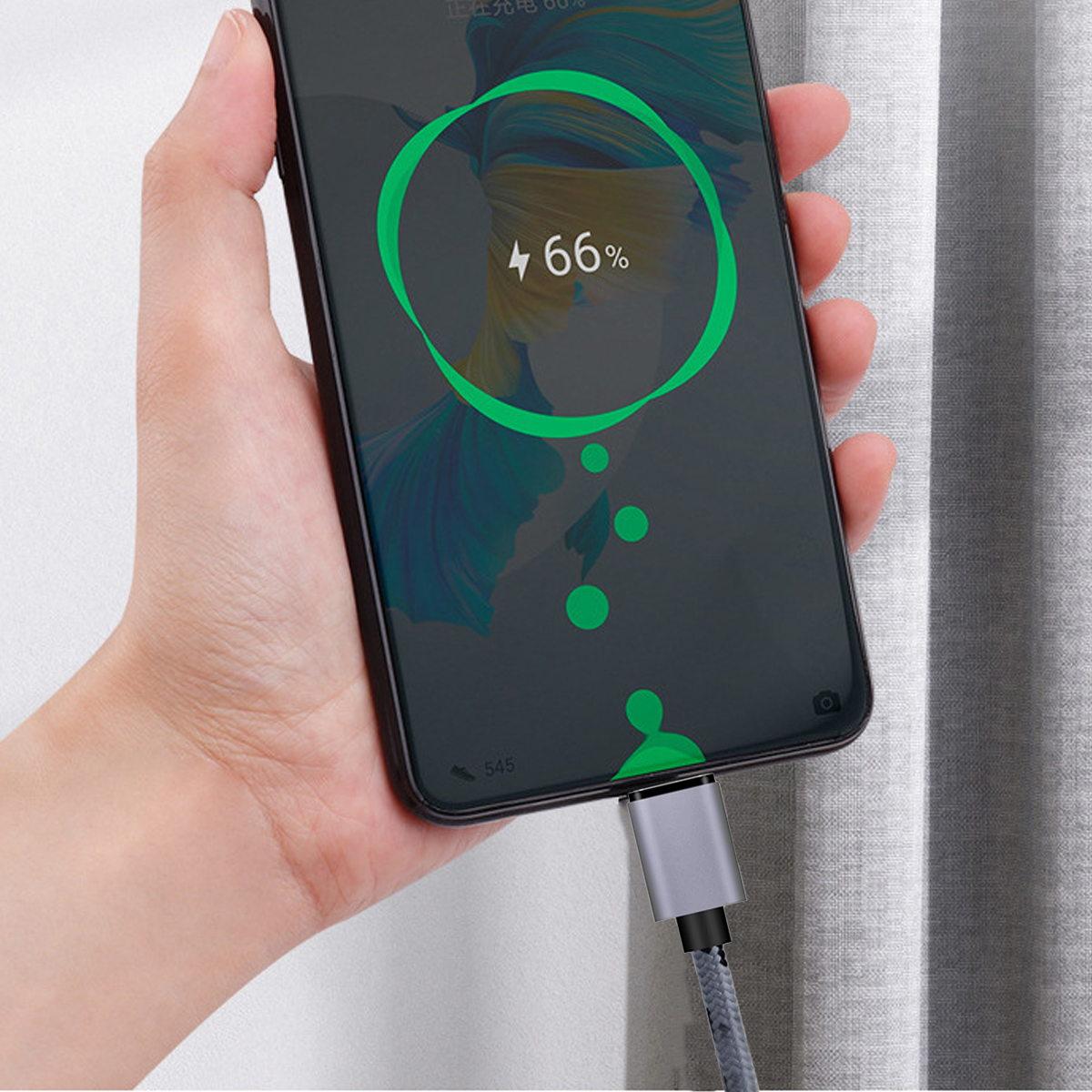Eine Person hält ein Telefon, das von einem grauen geflochtenen Kabel aufgeladen wird, das mit dem Ladeanschluss des Telefons verbunden ist.