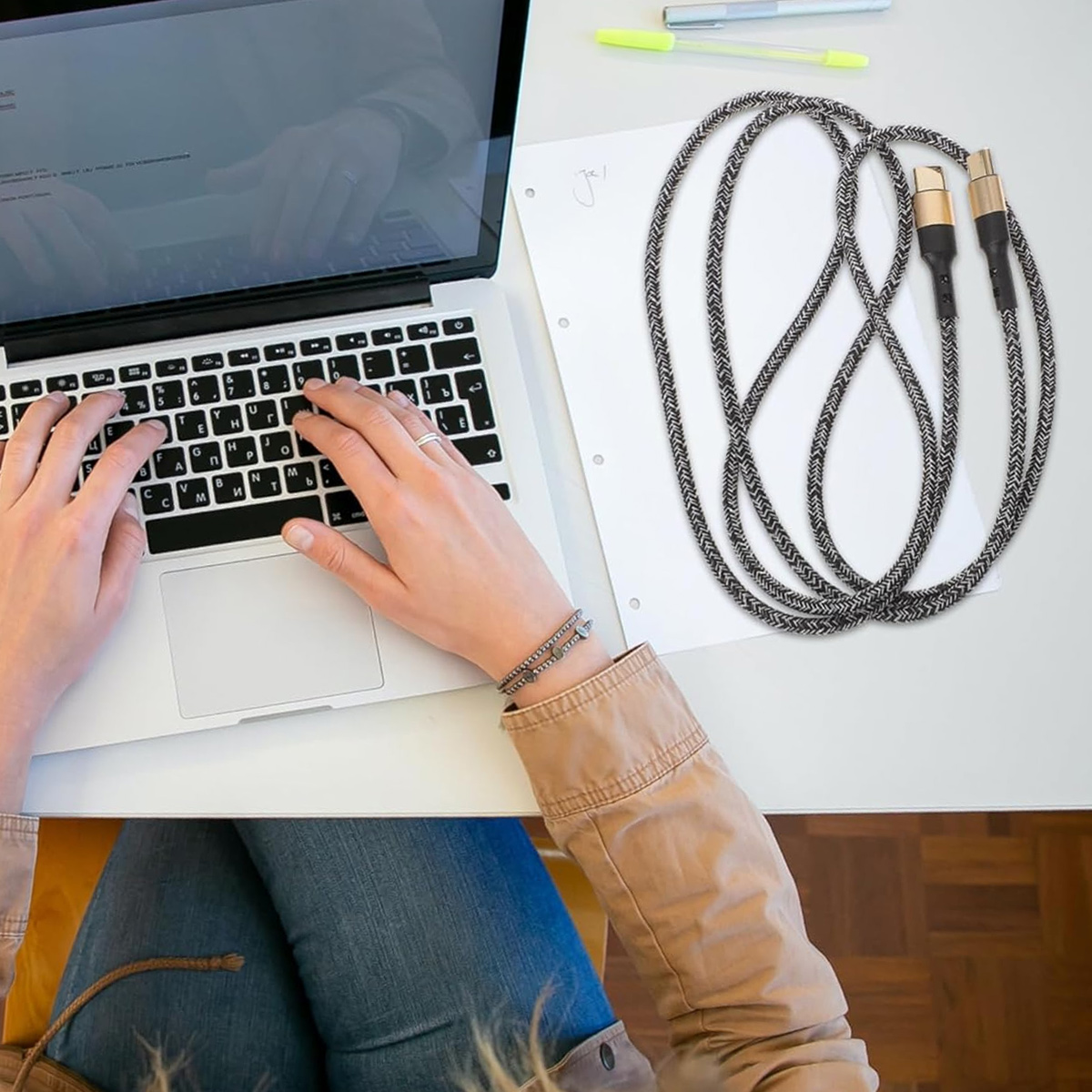 Person tippt auf einem Laptop, daneben liegt ein Kabel. Das Kabel ist schwarz und goldfarben. Weißer Hintergrund.