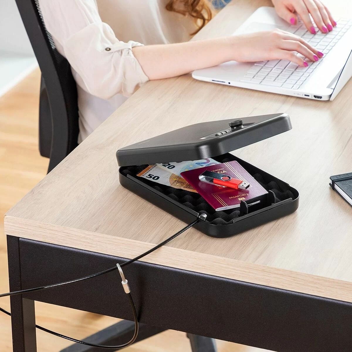 Una donna digita su un laptop accanto a una cassaforte nera aperta. All'interno denaro, passaporto e USB.