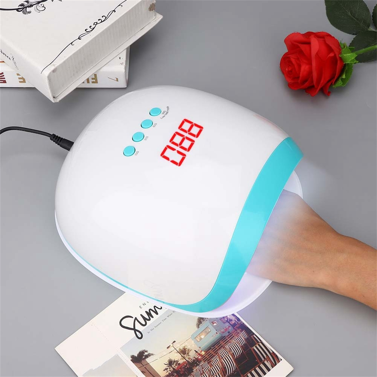 Eine Hand ist in einer Nagellampe. Die Lampe hat eine Digitalanzeige und ein blau-weißes Design. Eine Rose und ein Magazin sind in der Nähe.