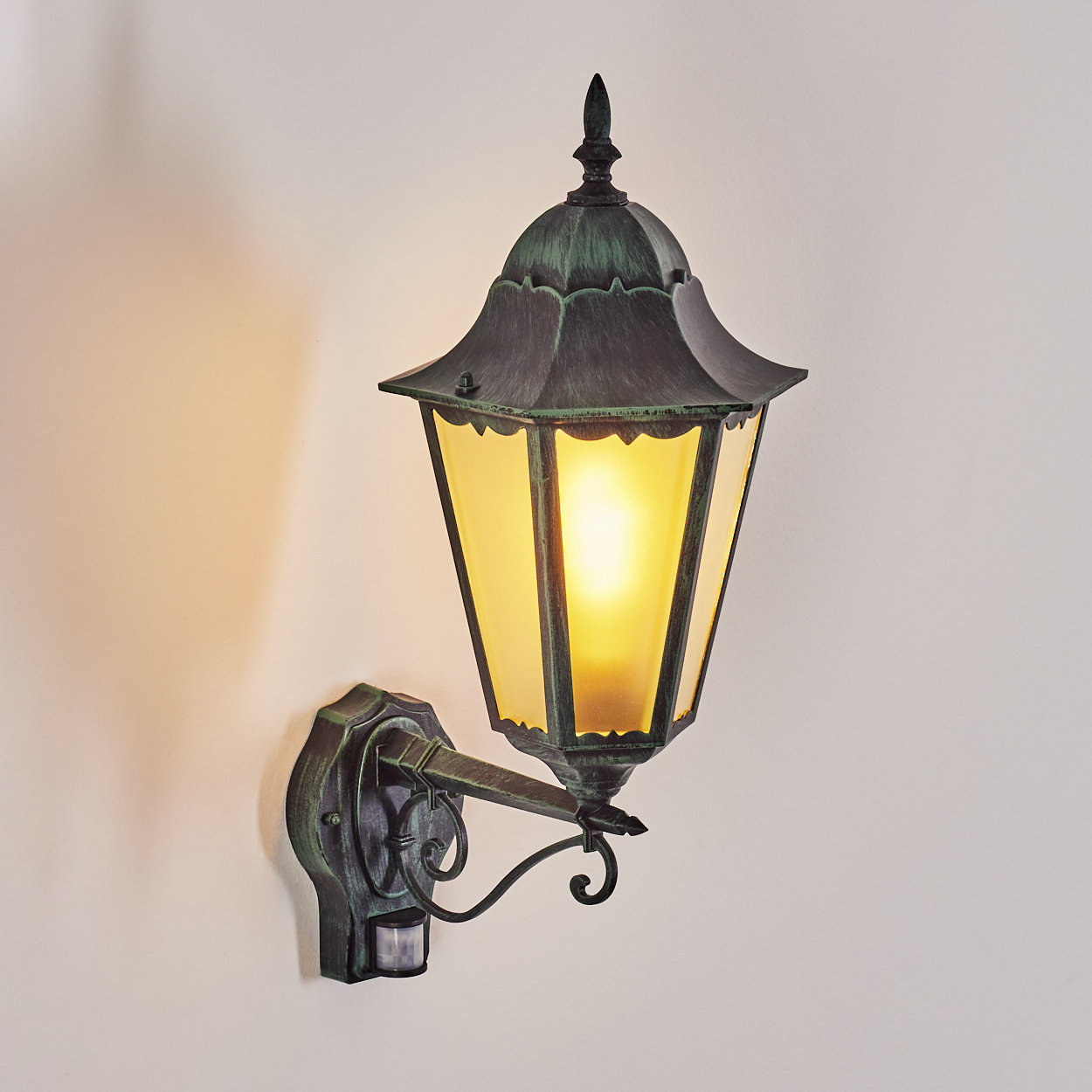 Außenleuchte an weißer Wand. Das Licht ist an und strahlt einen warmen gelben Schein aus. Bronzegrün.