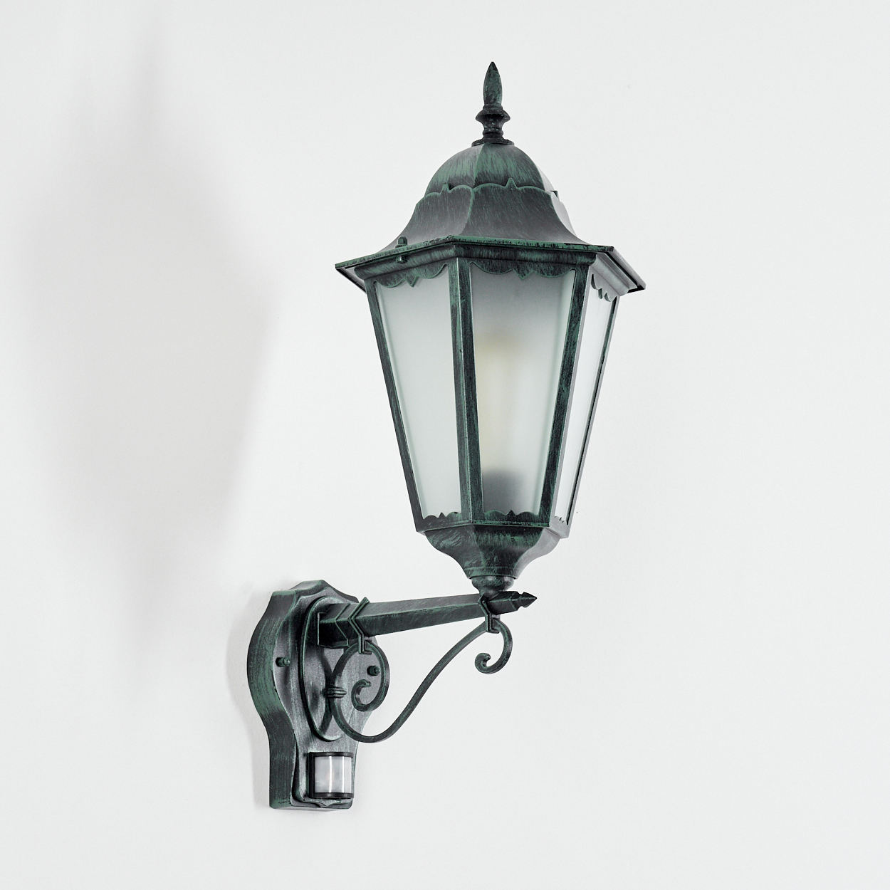 Eine kunstvolle, dunkelgrüne Außenleuchte an einer weißen Wand, die ein sanftes Licht ausstrahlt.