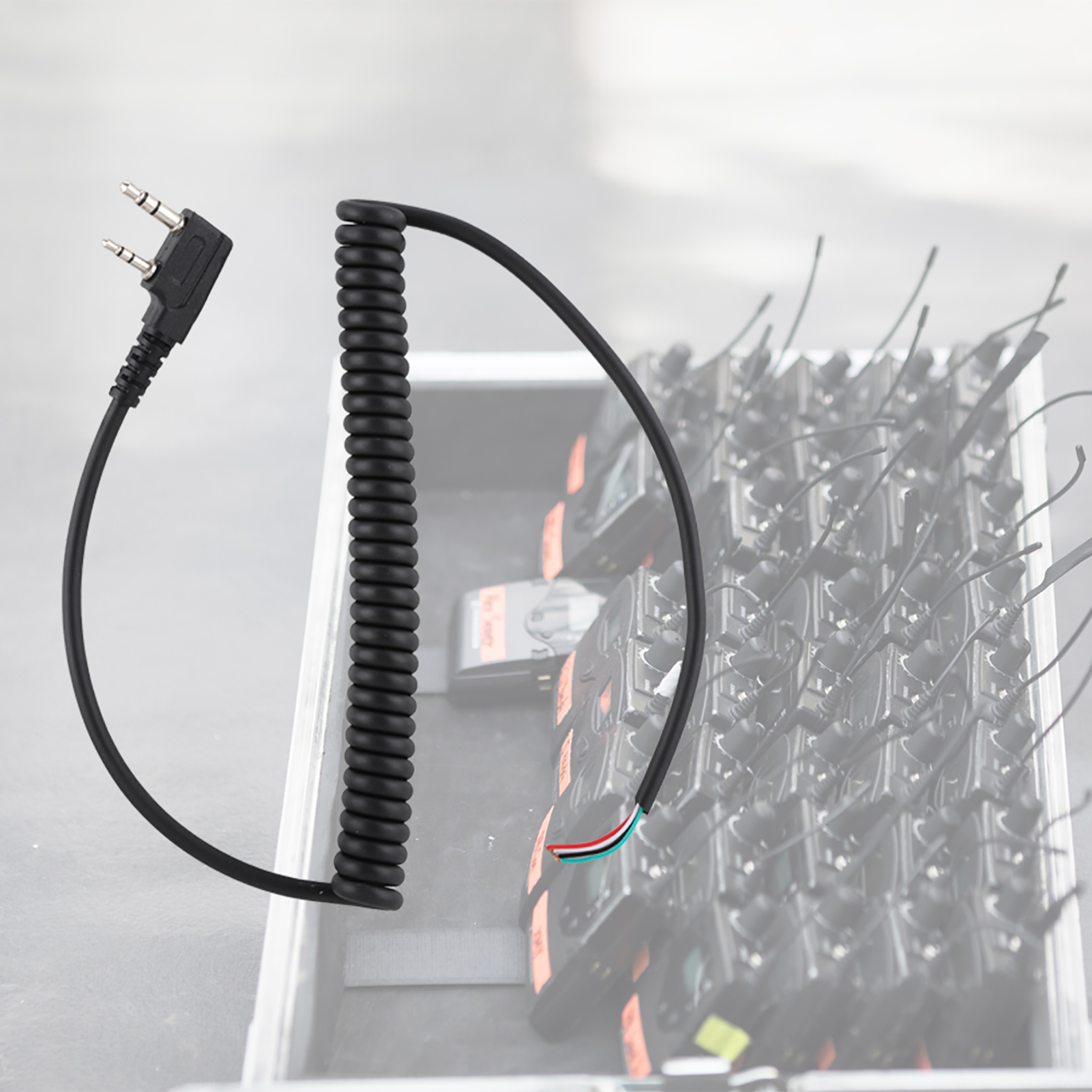 Ein schwarzes Spiralkabel mit einem 3,5-mm-Stecker und freiliegenden Drähten, mit Walkie-Talkies im Hintergrund.