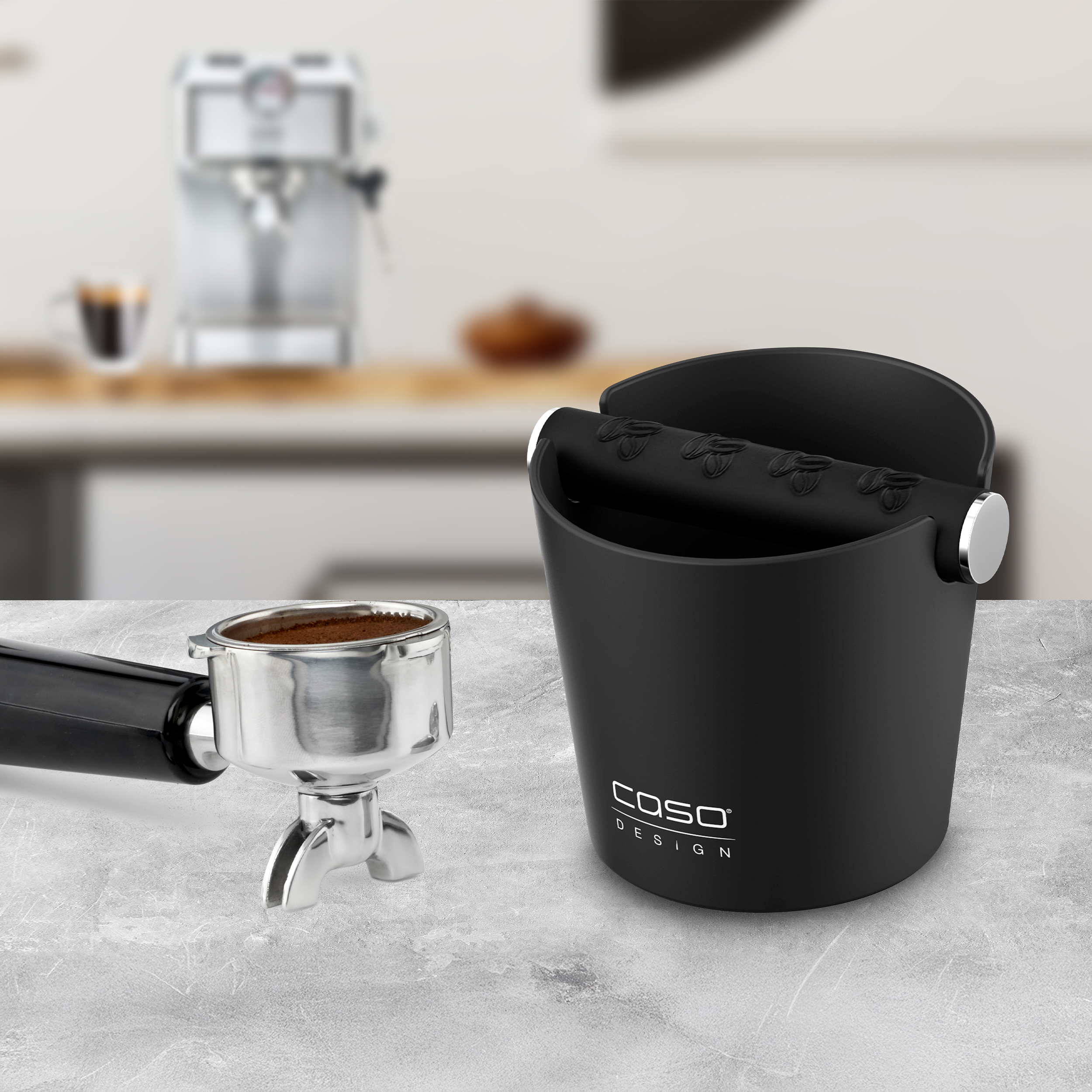 Ein Siebträger und eine schwarze Abschlagbox stehen auf einer grauen Oberfläche, im Hintergrund eine verschwommene Espressomaschine.