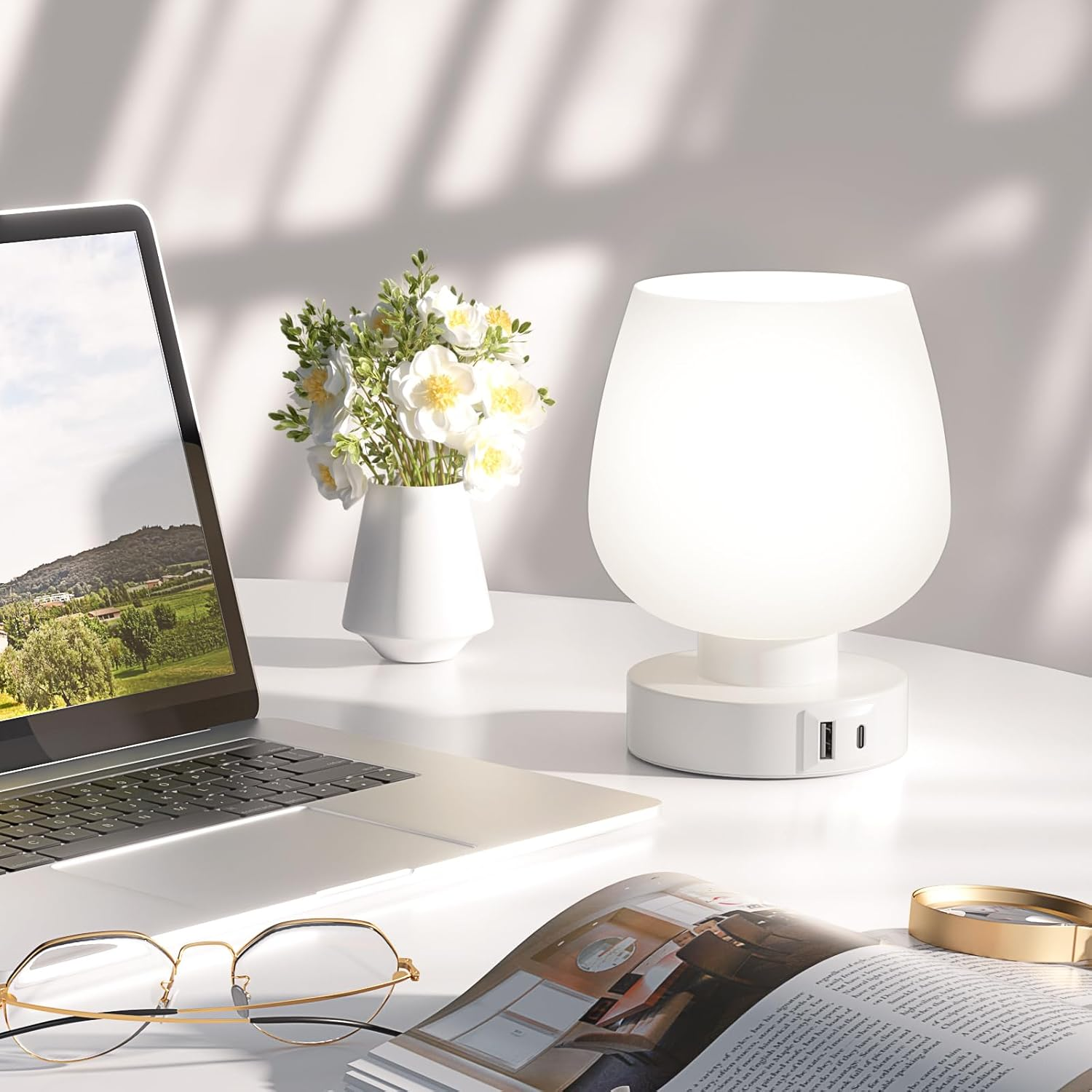 Eine weiße Touch-Lampe steht auf einem weißen Schreibtisch neben einem Laptop, Blumen, einer Brille und einer Zeitschrift.