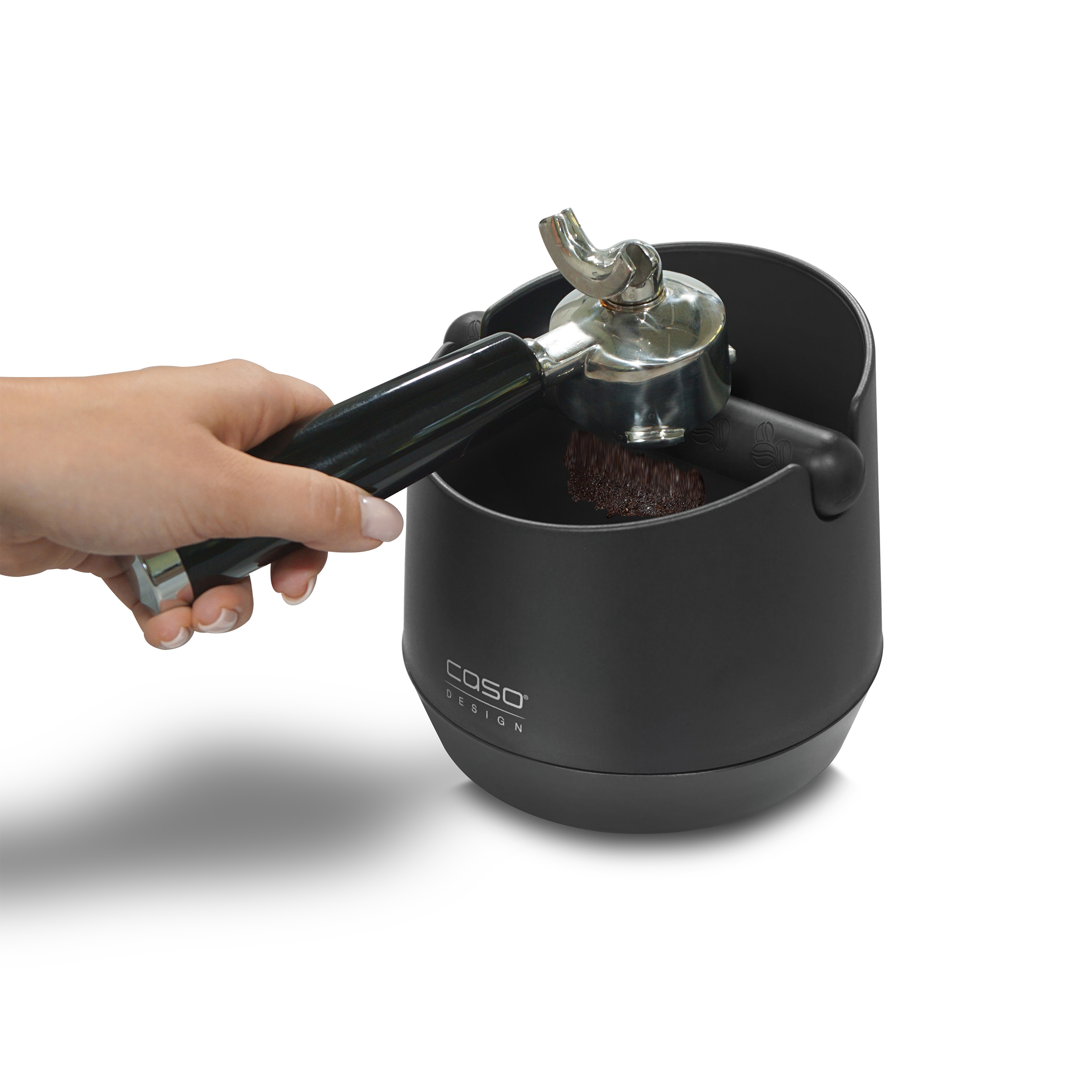 Eine Hand drückt Kaffeemehl in einen Siebträger über einer schwarzen Abschlagbox auf einer weißen Oberfläche.