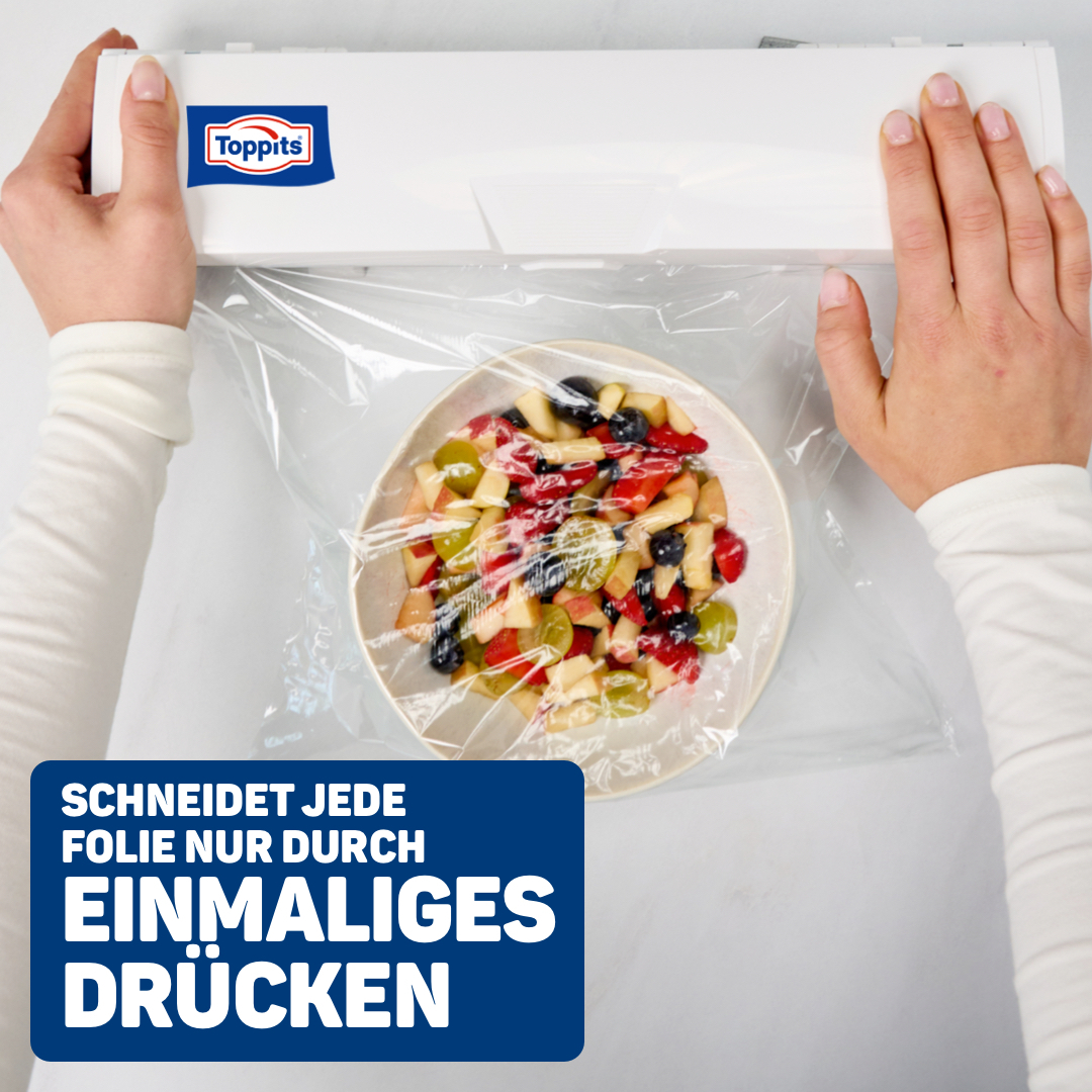 Hände spannen Plastikfolie über eine Schüssel mit Obstsalat, vor einem weißen Hintergrund.