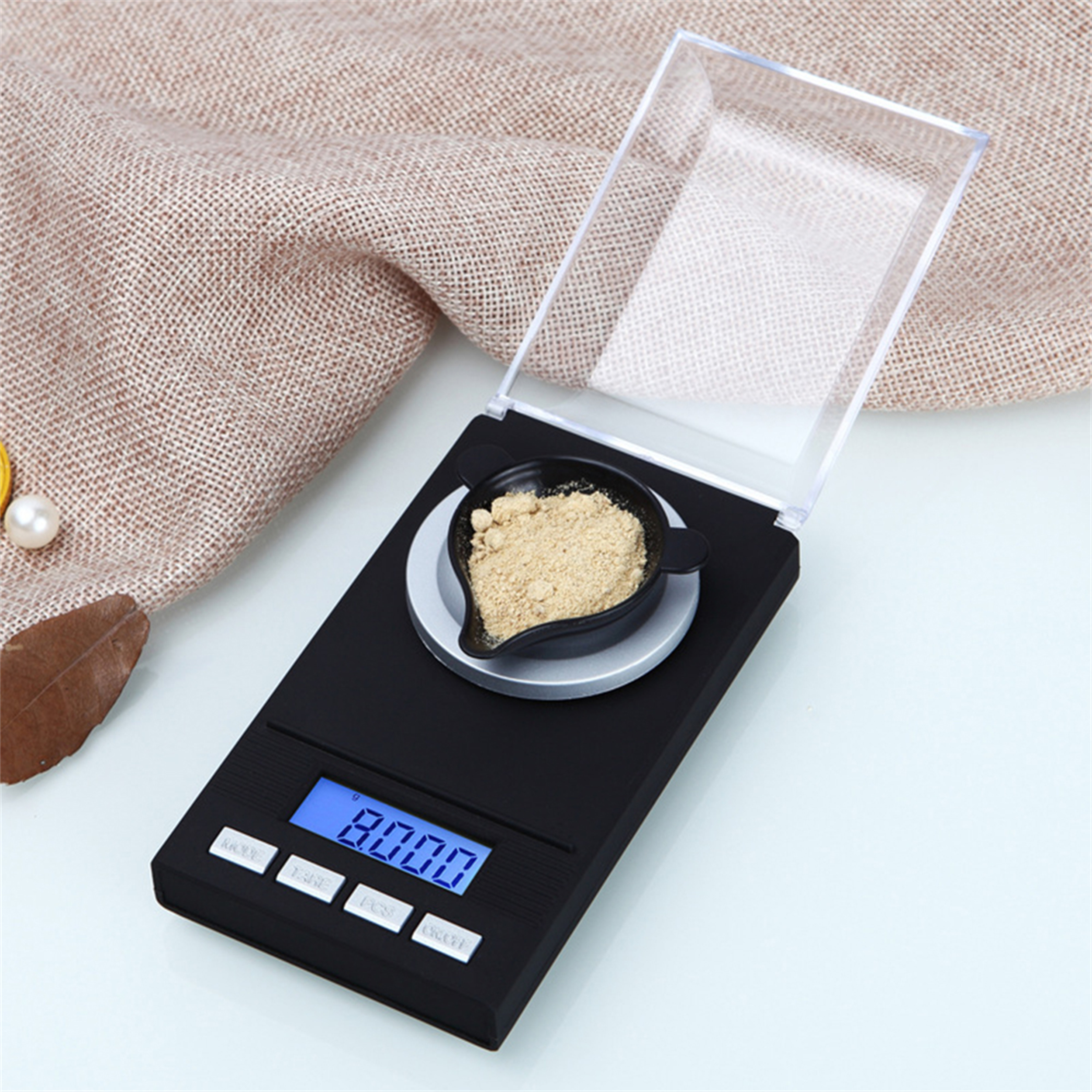 Eine schwarze Digitalwaage mit einem klaren Deckel, einer kleinen schwarzen Schale und etwas Pulver darin auf einer weißen Oberfläche.