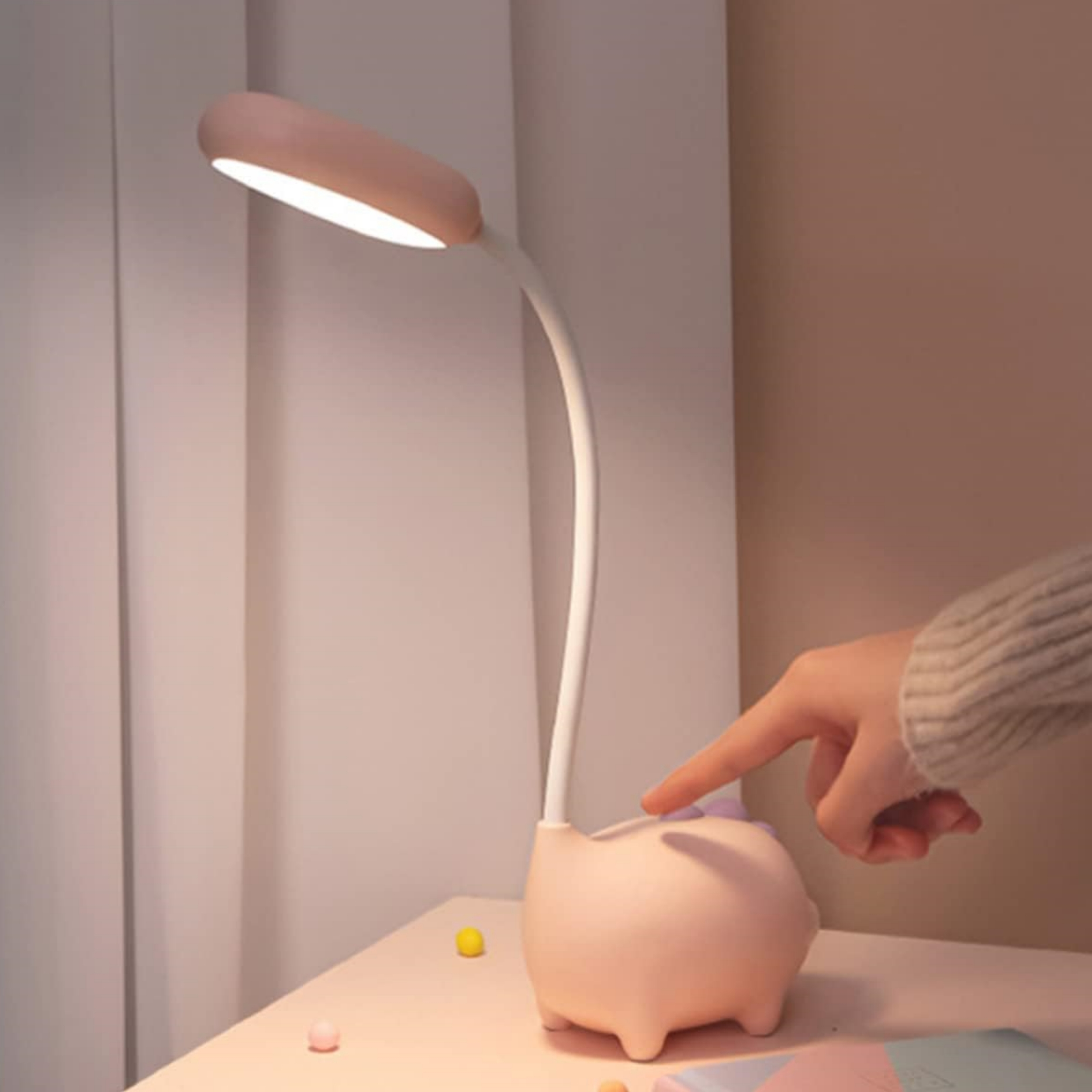 Eine Hand berührt eine rosa Schreibtischlampe in Schweineform. Sie steht auf einem Schreibtisch.