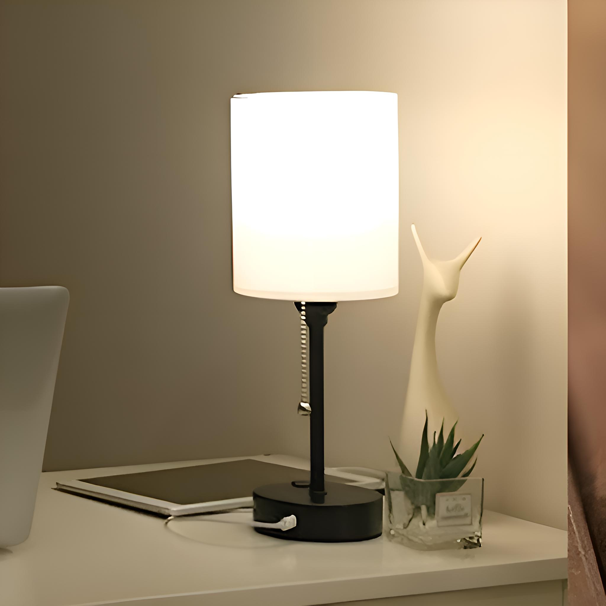 Eine beleuchtete weiße Lampe mit schwarzem Fuß steht auf einem weißen Schreibtisch neben einem Tablet, einer Sukkulente und moderner Deko.
