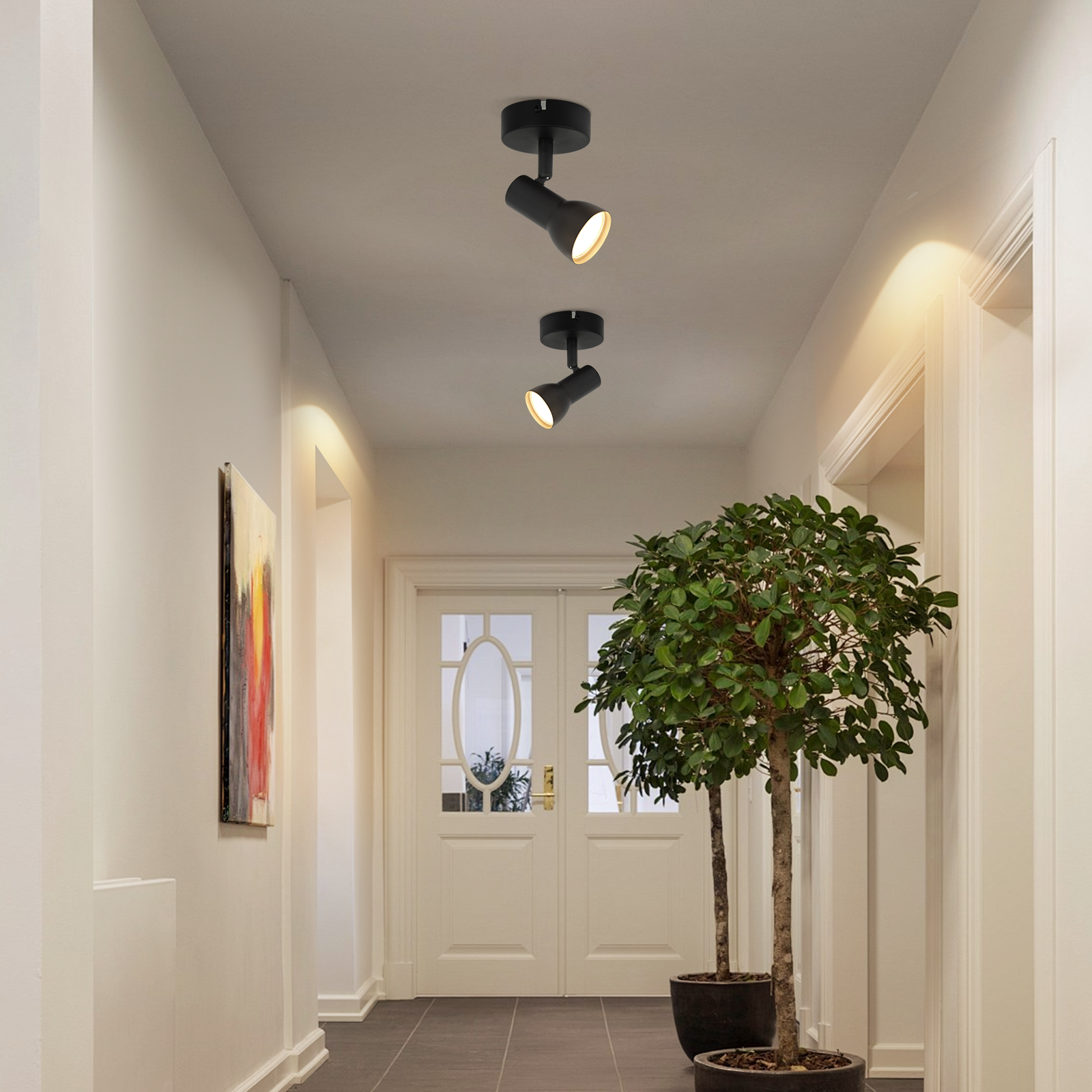 Dwa czarne reflektory sufitowe w korytarzu, drzewa w donicach i obraz na ścianie.