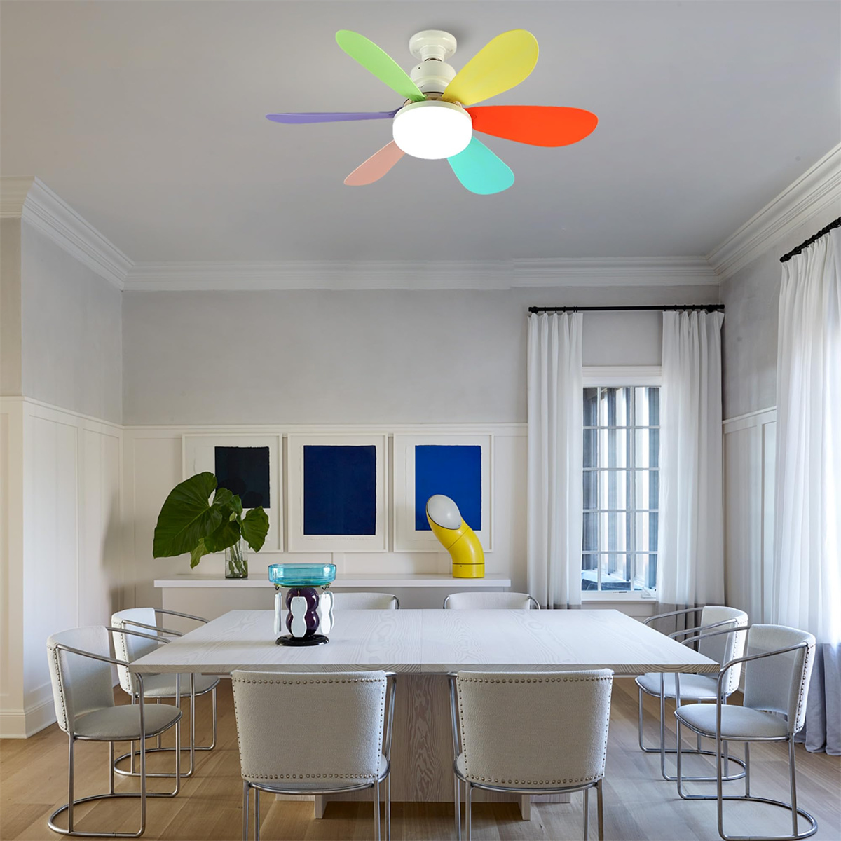 Ein Esszimmer mit einem weißen Tisch und Stühlen. Ein Deckenventilator mit bunten Flügeln hängt über dem Tisch.