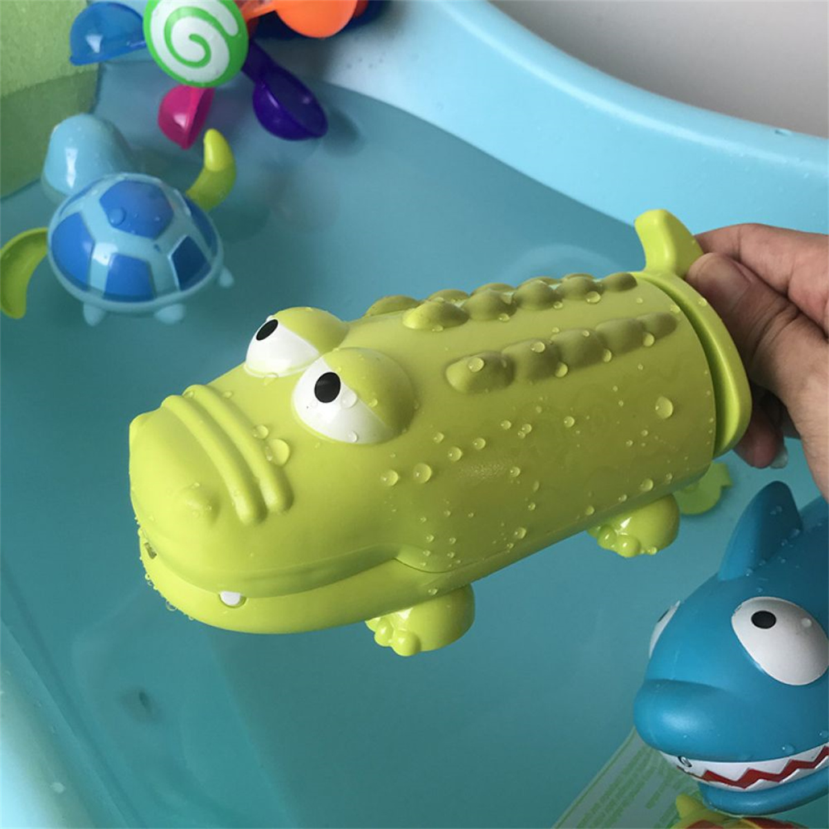 Eine grüne Krokodil-Wasserpistole wird in der Hand gehalten, mit Badespielzeug im Hintergrund.