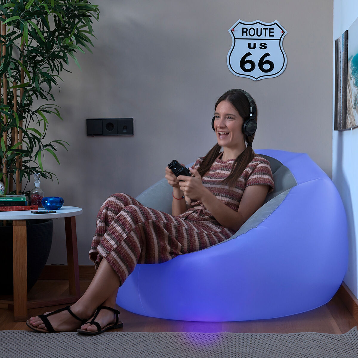 Eine lächelnde Frau sitzt in einem blauen aufblasbaren Stuhl und spielt mit einem Controller in einem Raum mit Pflanzen und einem Route 66-Schild.