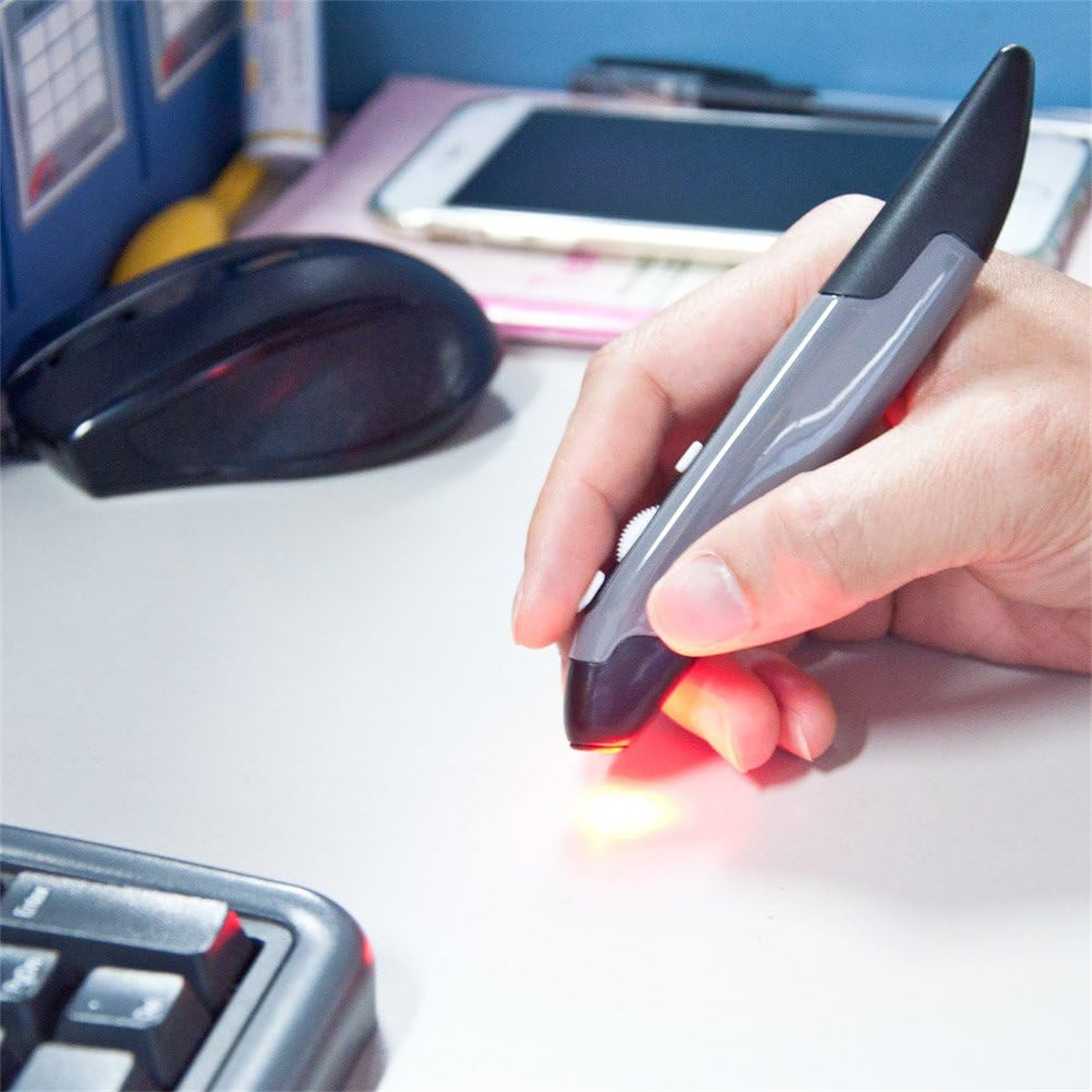 Eine Hand hält eine graue Stiftmaus mit rotem Licht, Tastatur, Maus, Telefon und Akten liegen auf einem weißen Schreibtisch.