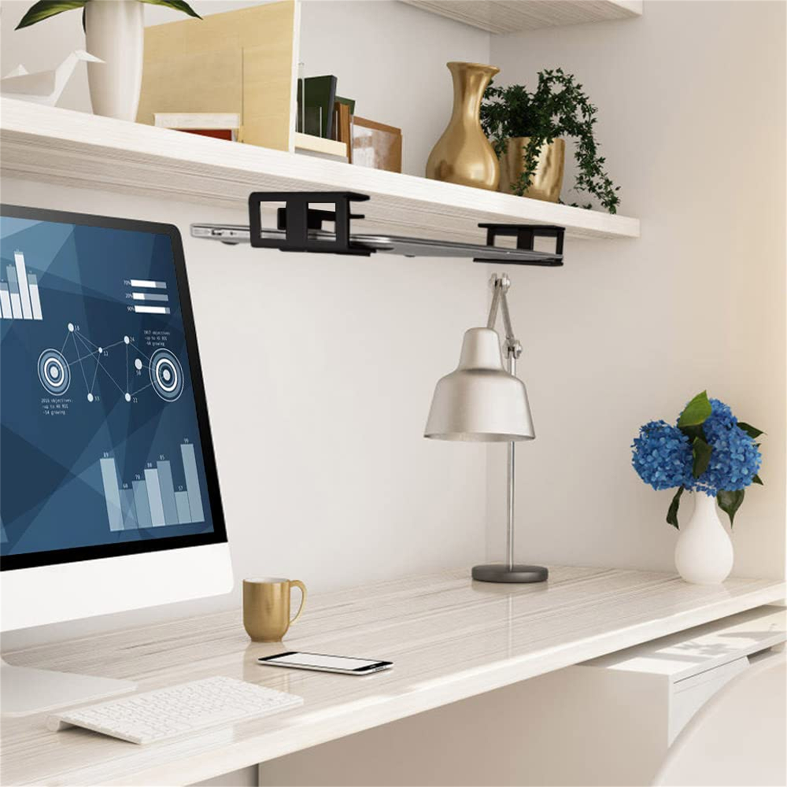 Ein Schreibtisch mit Monitor, Tastatur, Lampe und Blumen. Ein Regal darüber hält einen Computerständer.