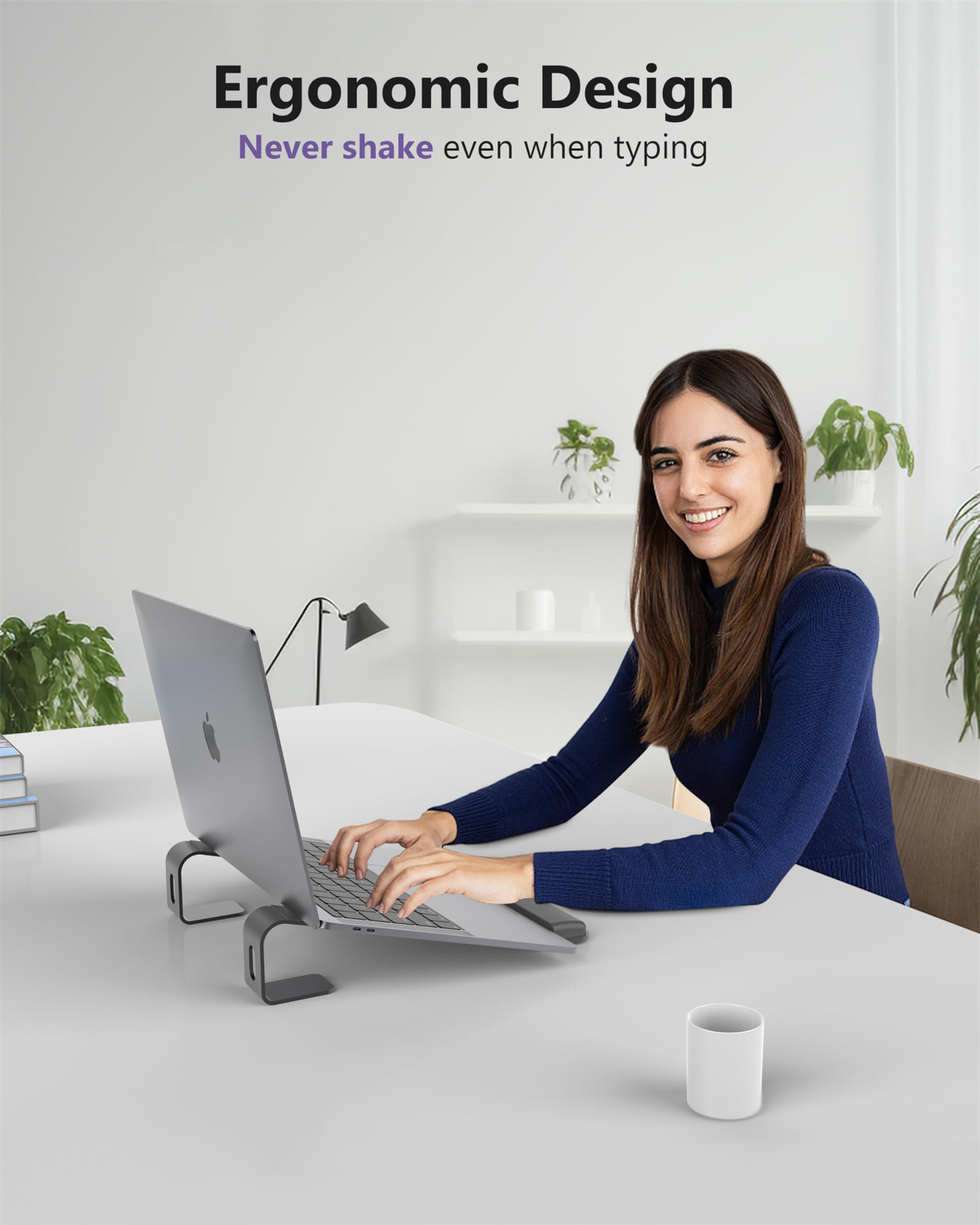 Frau sitzt mit Laptop auf einem grauen Ständer am Schreibtisch. Sie lächelt und hat eine Tasse auf dem Schreibtisch.