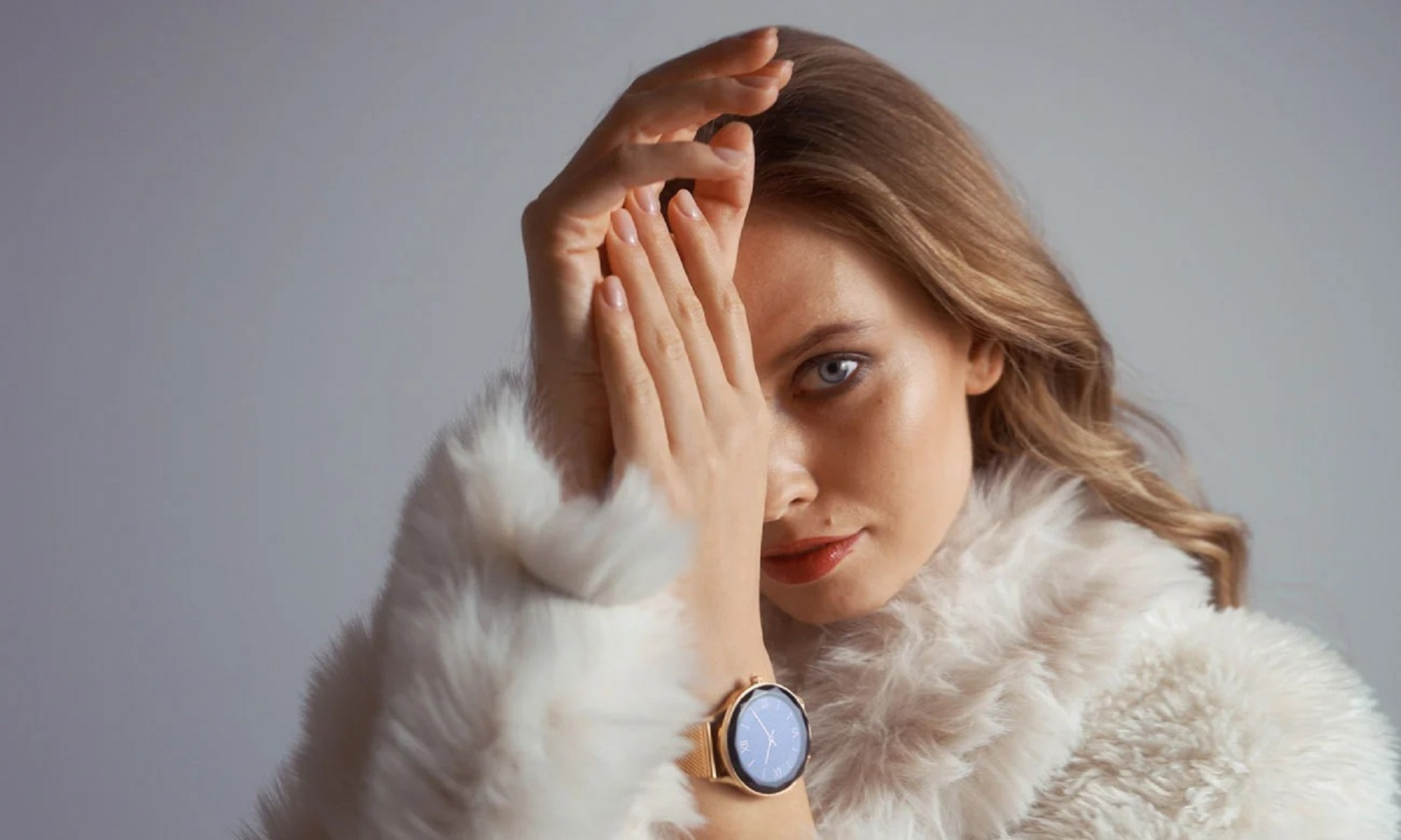Eine Frau im Pelzmantel bedeckt ihr Gesicht mit der Hand und trägt eine roségoldene Smartwatch.