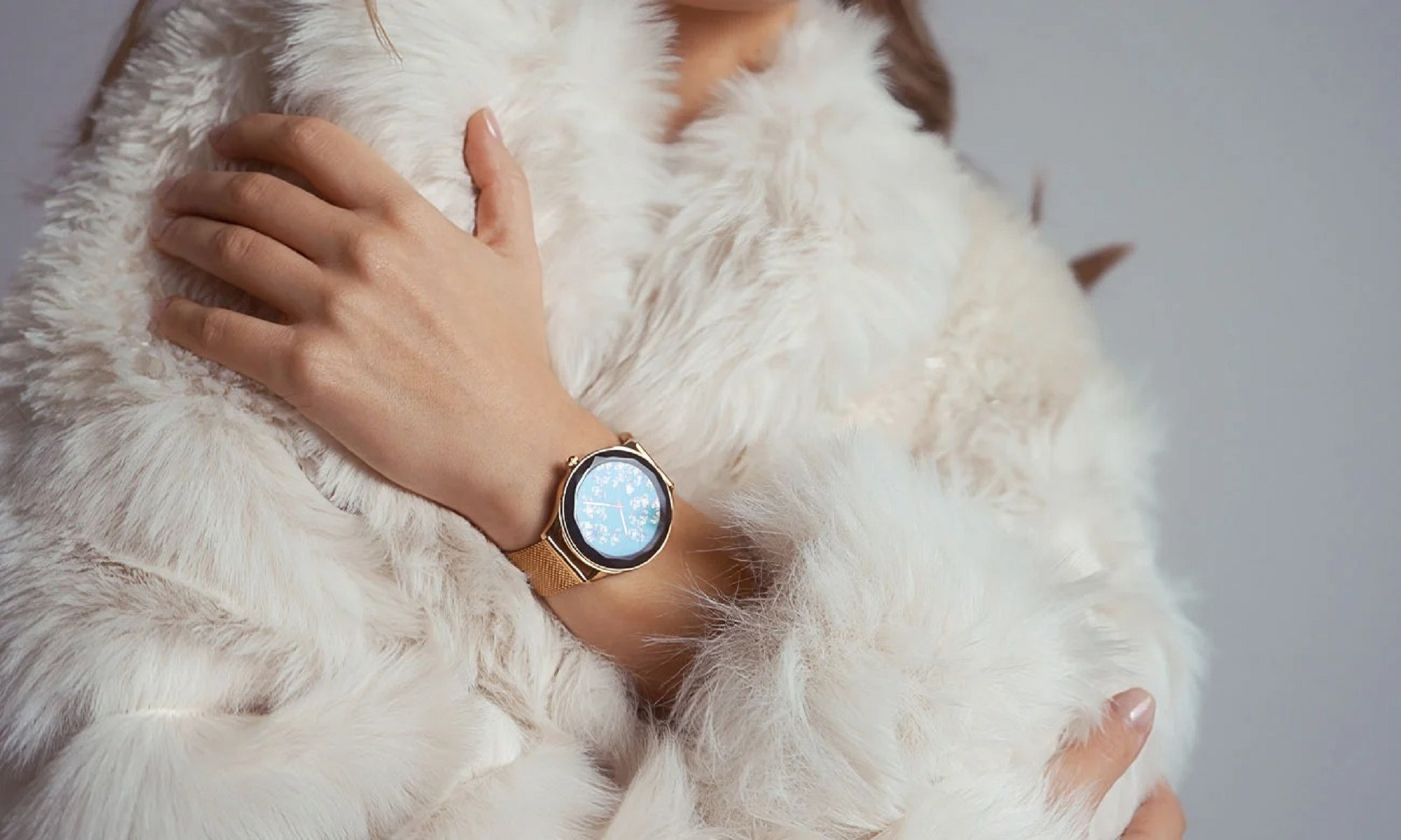 Nahaufnahme des Arms einer Frau, die in einem weißen Pelzmantel eine roségoldene Smartwatch mit Mesh-Armband trägt.