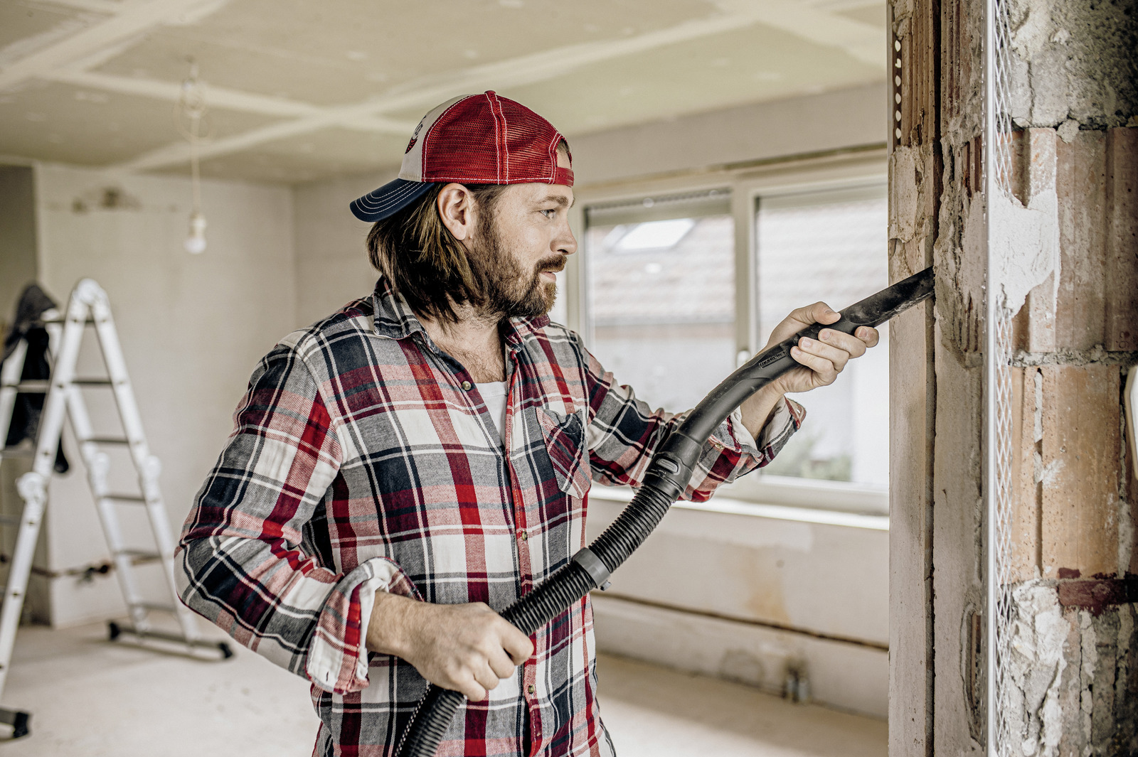 Mężczyzna w czapce używa odkurzacza w pokoju z odsłoniętymi ścianami.