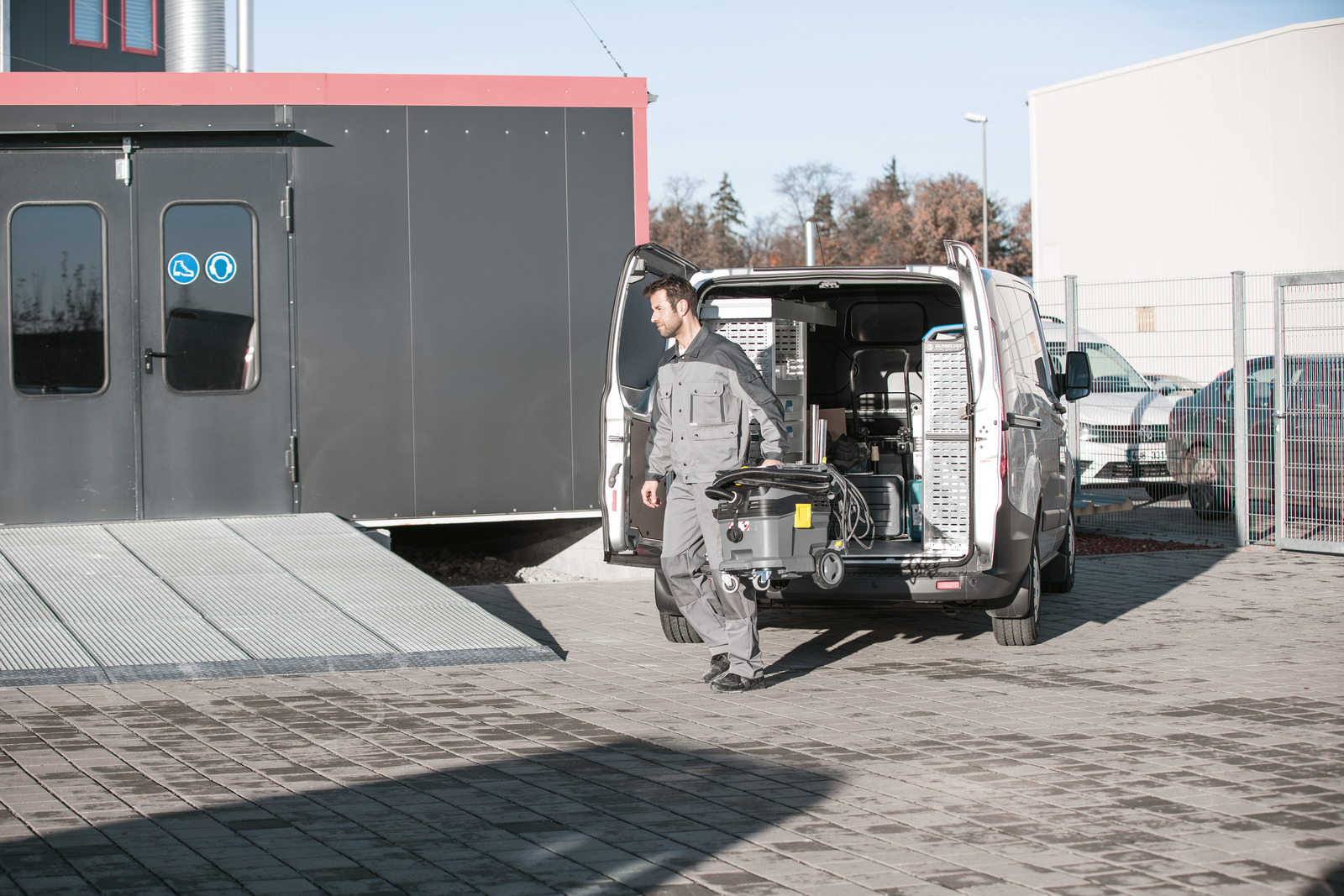 Ein Mann lädt einen grauen Staubsauger aus einem grauen Van auf einem Parkplatz aus.