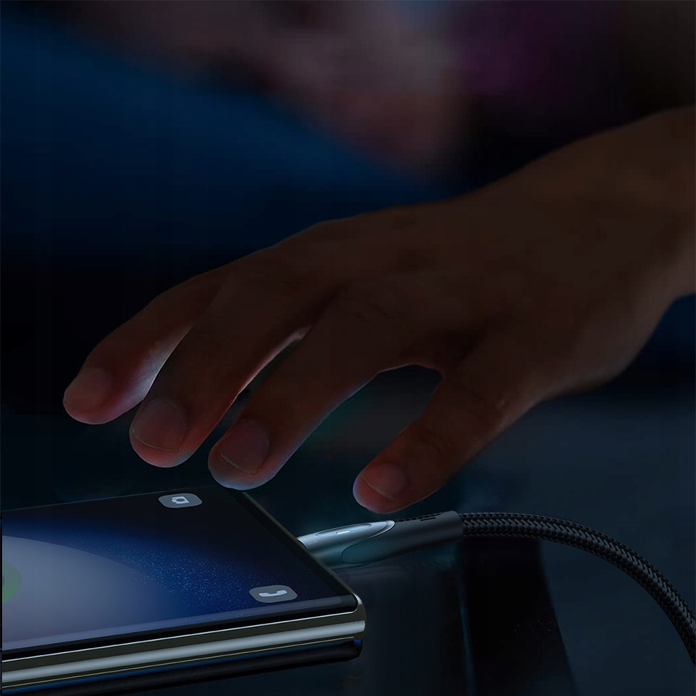 Ręka sięga po telefon ładujący się za pomocą plecionego czarnego kabla w słabo oświetlonym pomieszczeniu.