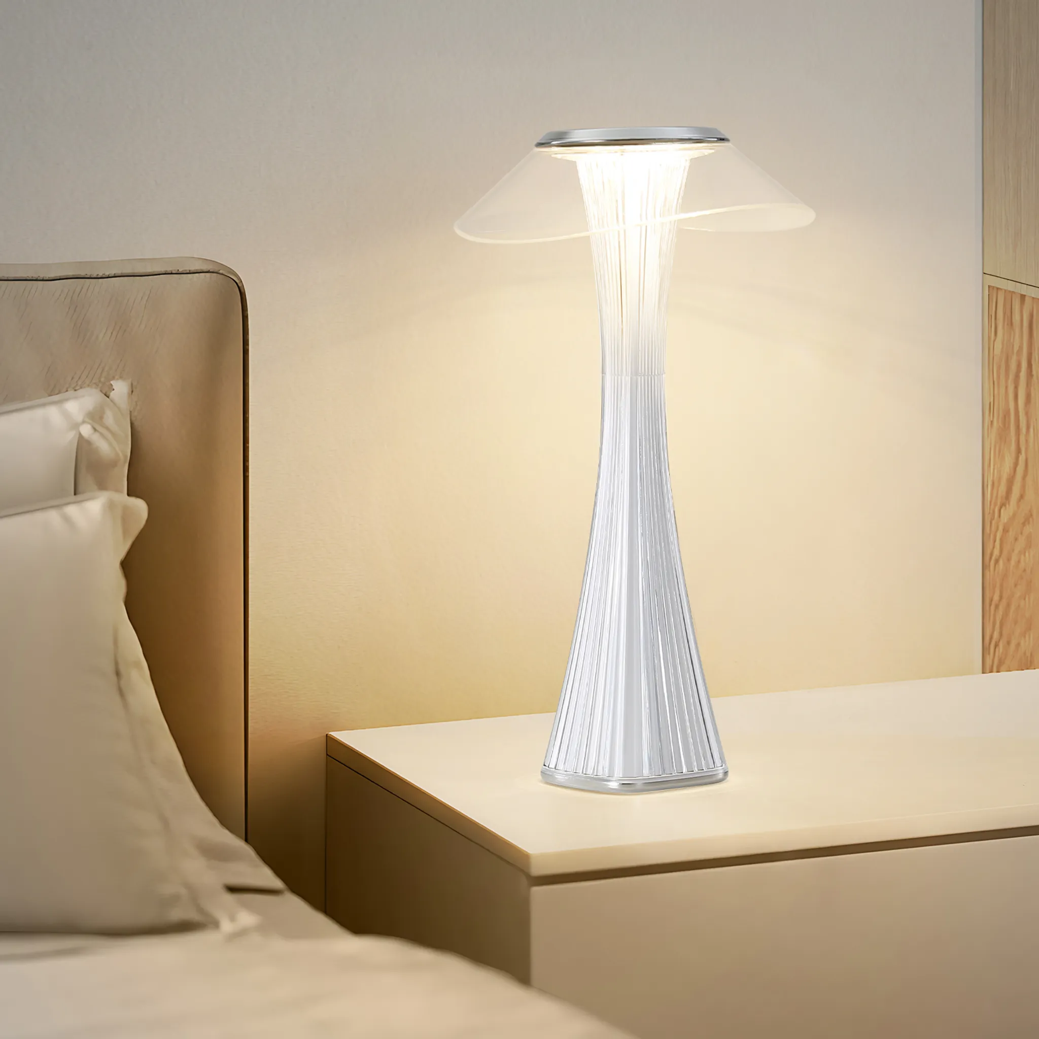 Biała lampa z przezroczystym kloszem stoi na białej szafce nocnej. Lampa jest włączona, emitując ciepłe, miękkie światło.