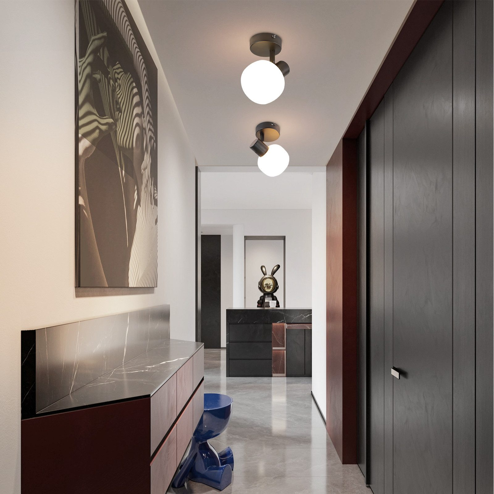 Un corridoio con due lampade a soffitto. Il corridoio ha una porta in legno scuro e un dipinto sulla parete sinistra.
