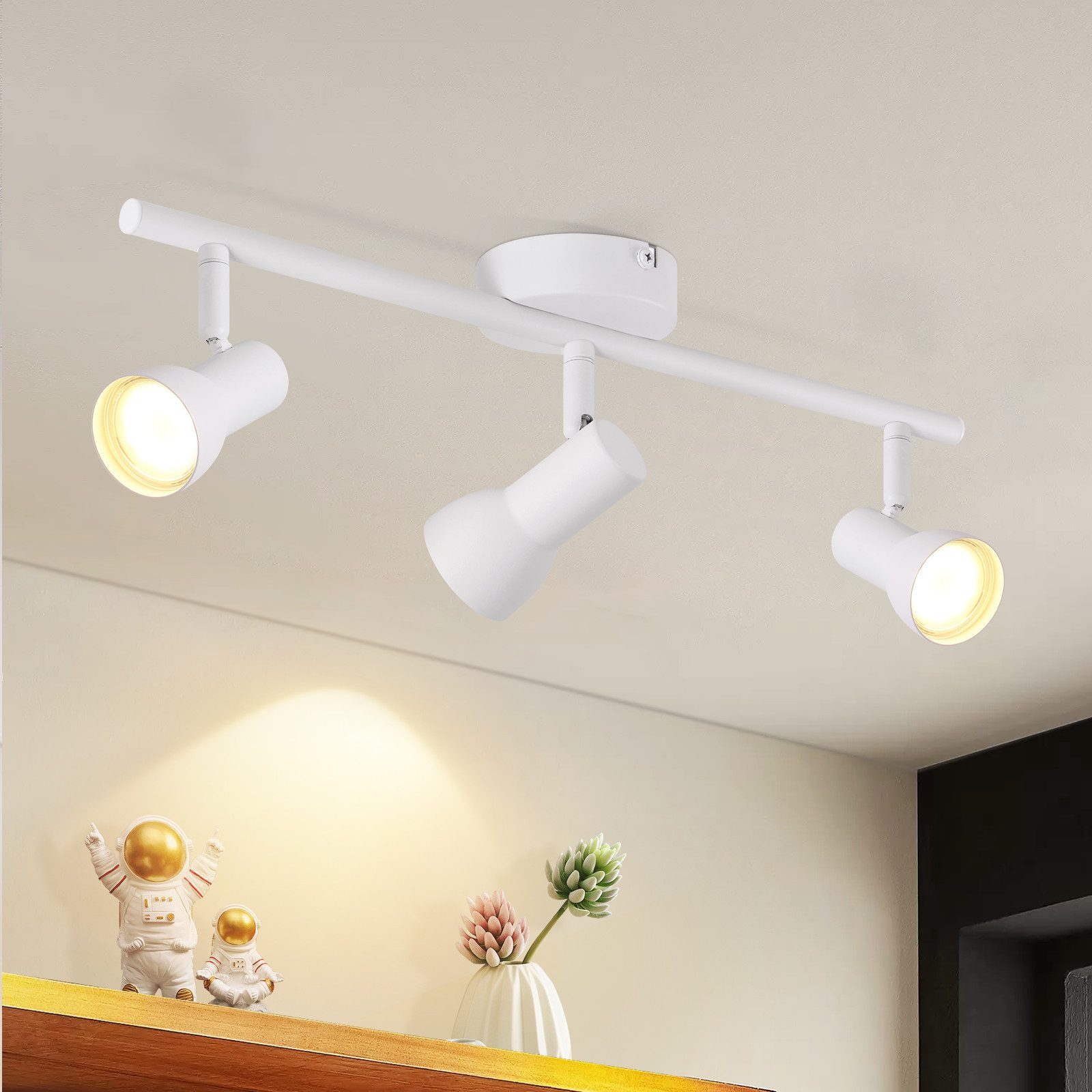Plafoniera bianca con tre faretti regolabili. Oggetti decorativi sono sullo scaffale sottostante. Lo sfondo è sfocato.