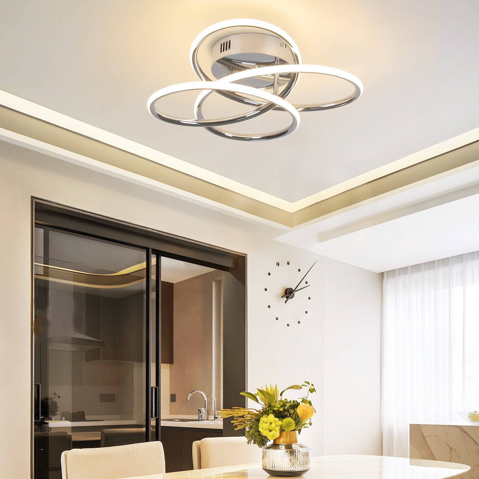 Un comedor moderno con una lámpara de techo LED cromada, una mesa de cristal con flores y una puerta corredera negra a una cocina.