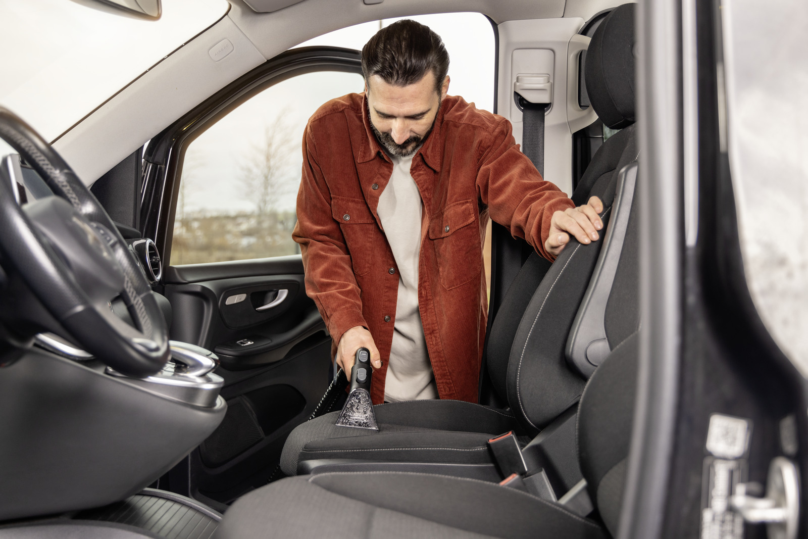 A man in an orange shirt cleans a car seat with a handheld vacuum cleaner, focusing on detailed cleaning.