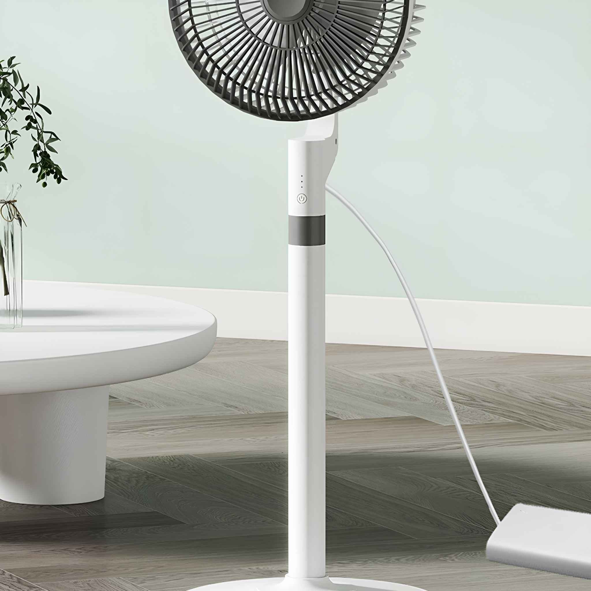 Ein stehender Ventilator mit weißem Gehäuse und grauem Lüftergitter ist neben einem weißen Tisch und einer grünen Pflanze zu sehen.