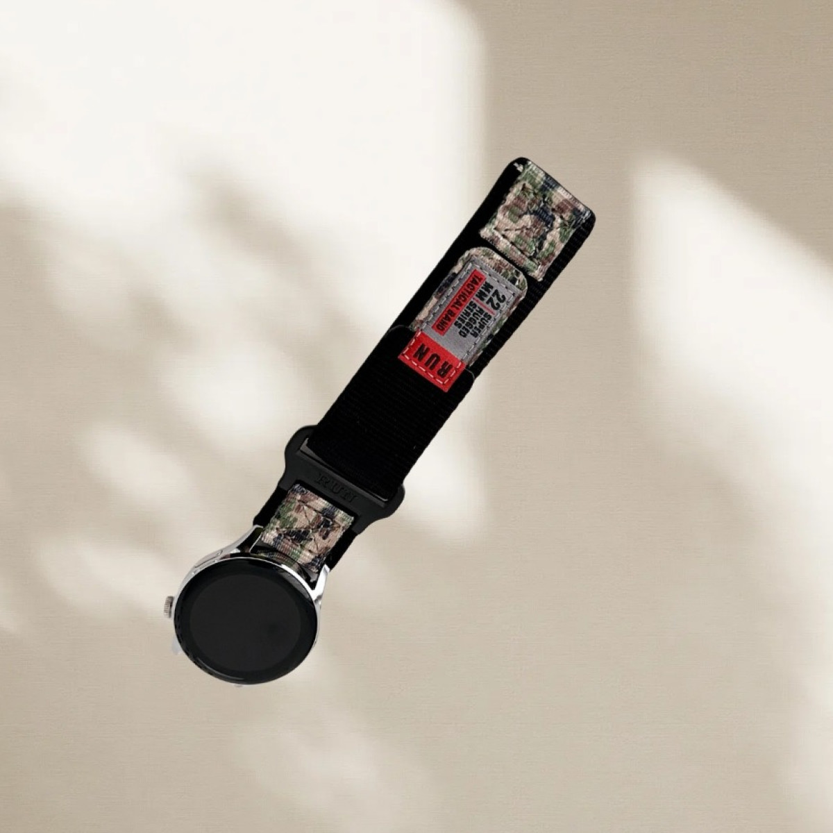 Uno smartwatch con cinturino tattico in nylon nero, con inserti mimetici e un'etichetta rossa con la scritta 'RUN', angolato su una superficie beige.